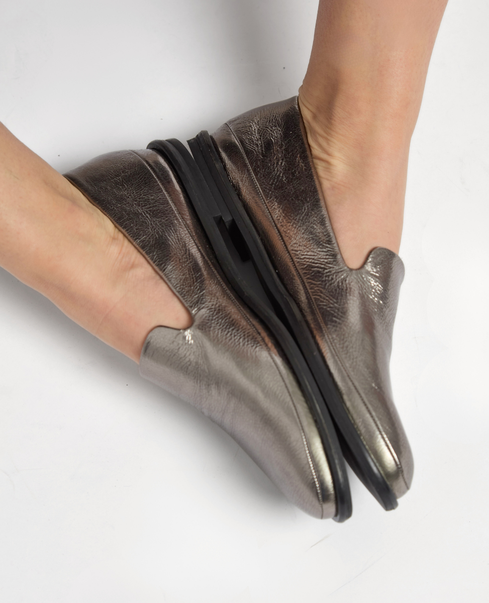 Metallic silver slip-on shoes with a smooth, shiny texture, featuring a slightly pointed toe and a black, flat sole, designed to be flexible and foldable.