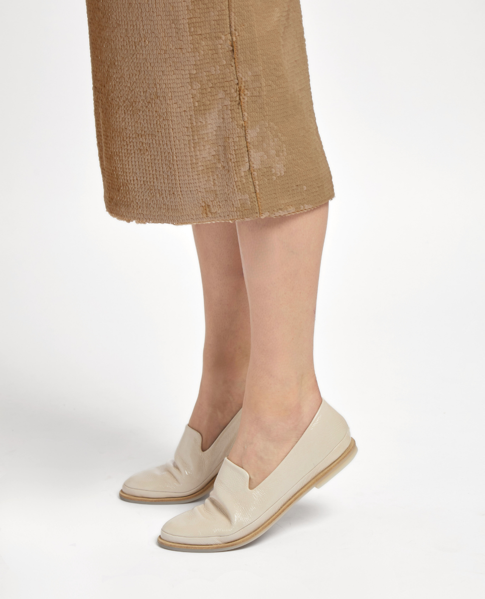 A pair of sleek, cream-colored loafers with a glossy finish, featuring a low heel and a smooth, rounded toe design, worn with a textured, light brown skirt.