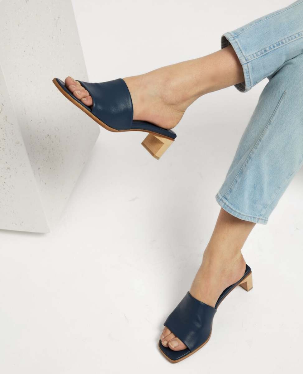 A pair of navy blue, open-toe mule sandals with a wide leather strap, square toe, and a low, light wooden block heel, worn by a person in light blue jeans.