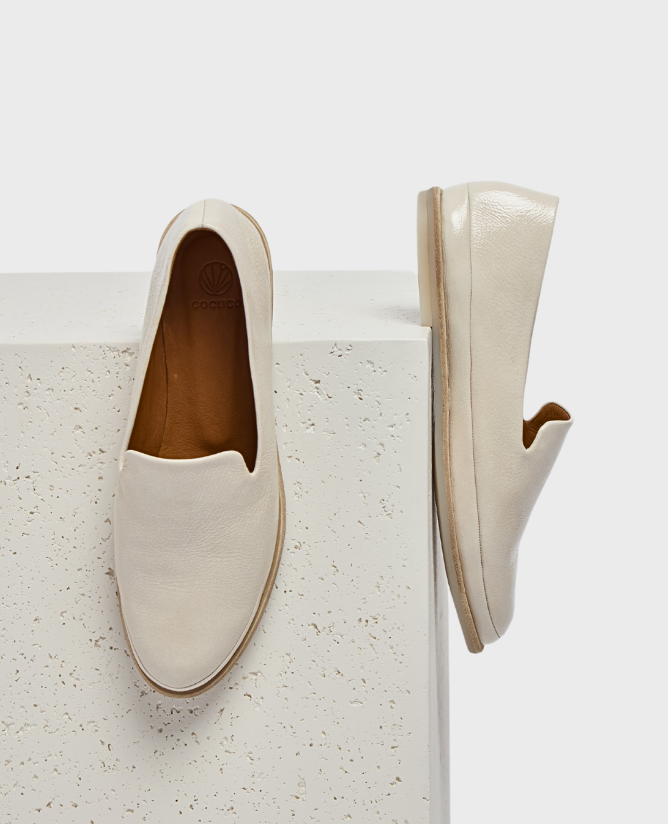 A pair of sleek, cream-colored slip-on shoes with a smooth leather texture, featuring a rounded toe, a low-profile rubber sole, and a subtle logo embossed on the insole.