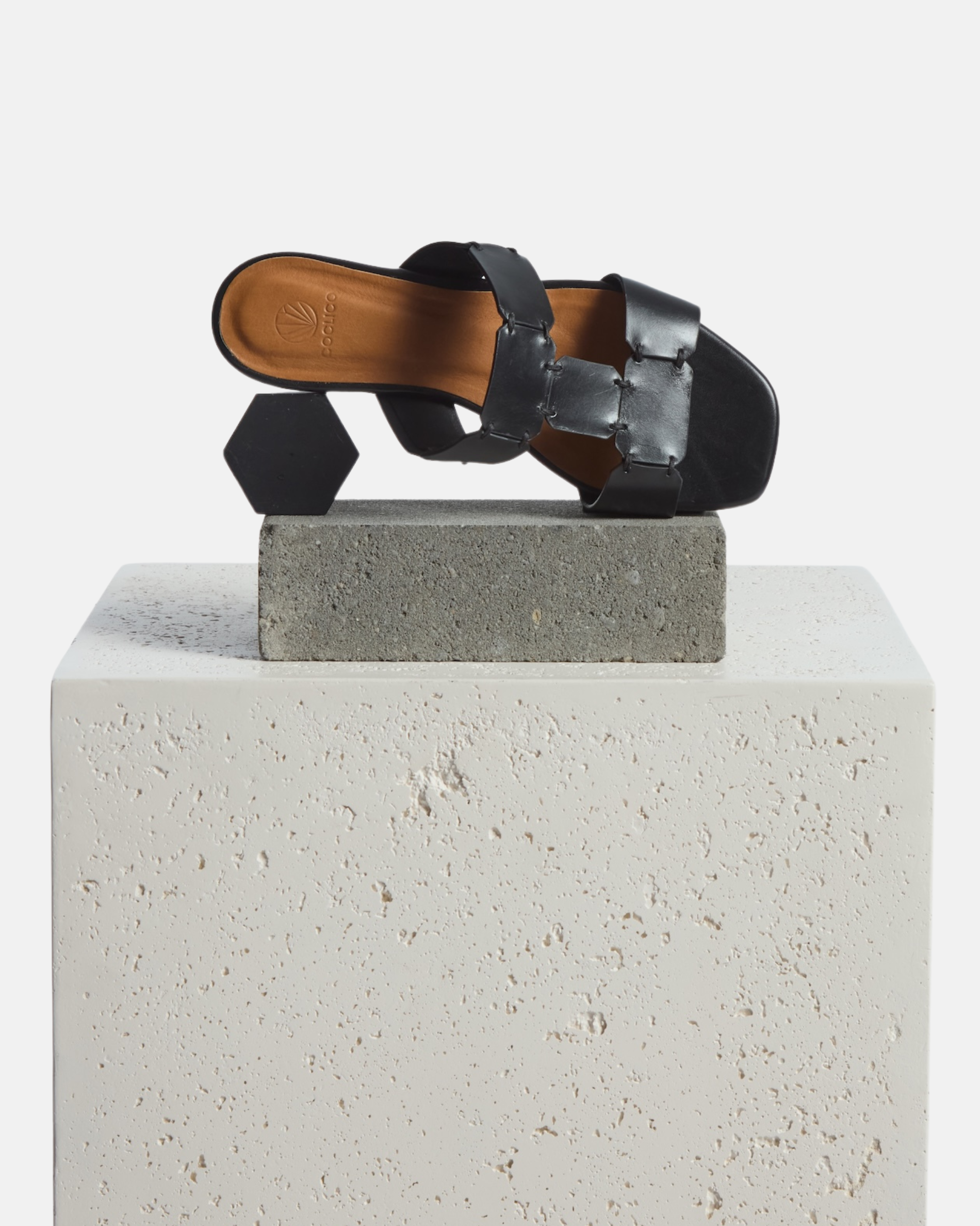 Shoe on Plinth: black vachetta leather T-strap slide sandal with octagonal hand stitched panels, square toe, and flat sole, on a white background.