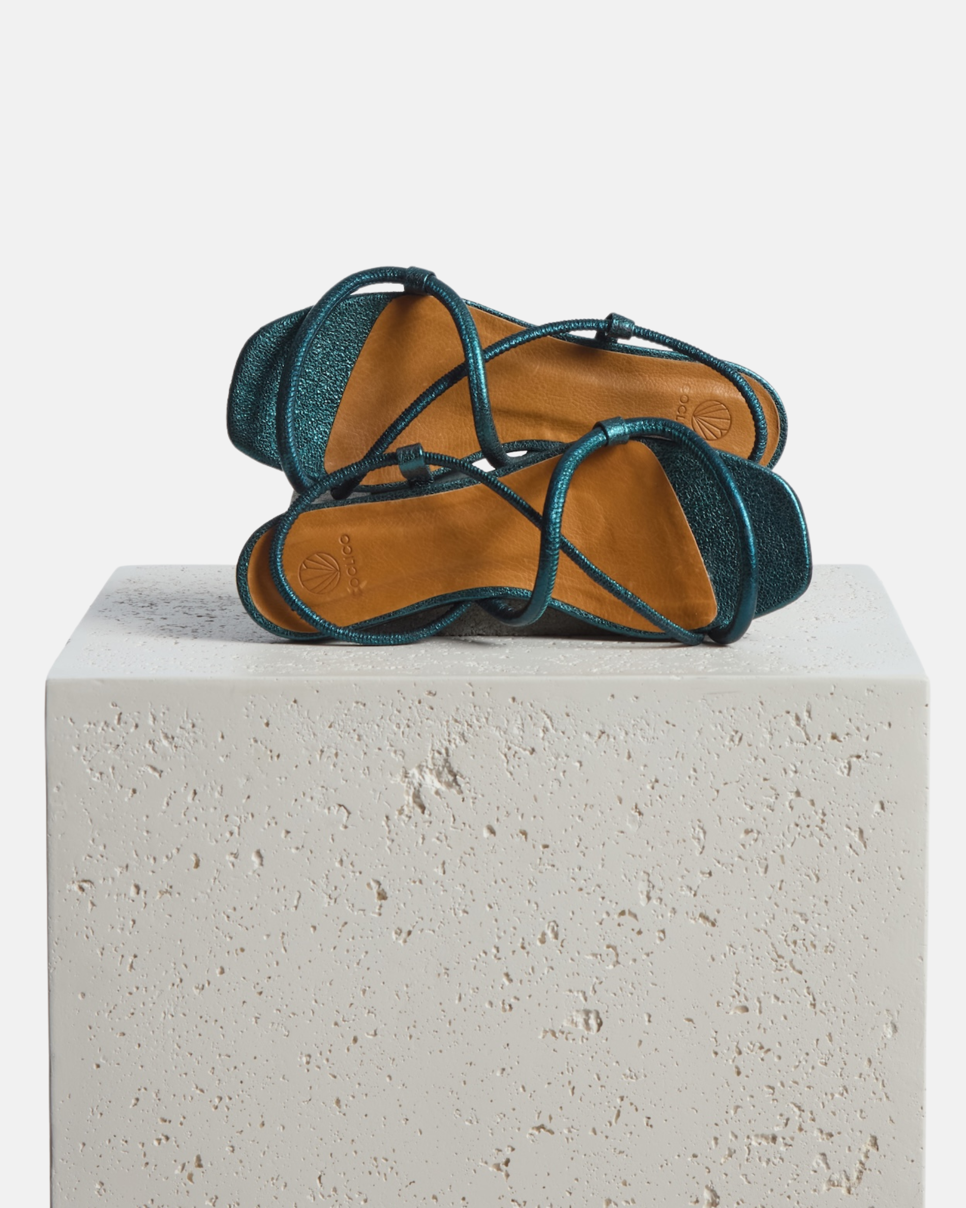 Shoes arranged on a stone pedestal: metallic blue crinkled leather flat sandal with slim padded straps, elasticated slingback, and a square toe on a grey background