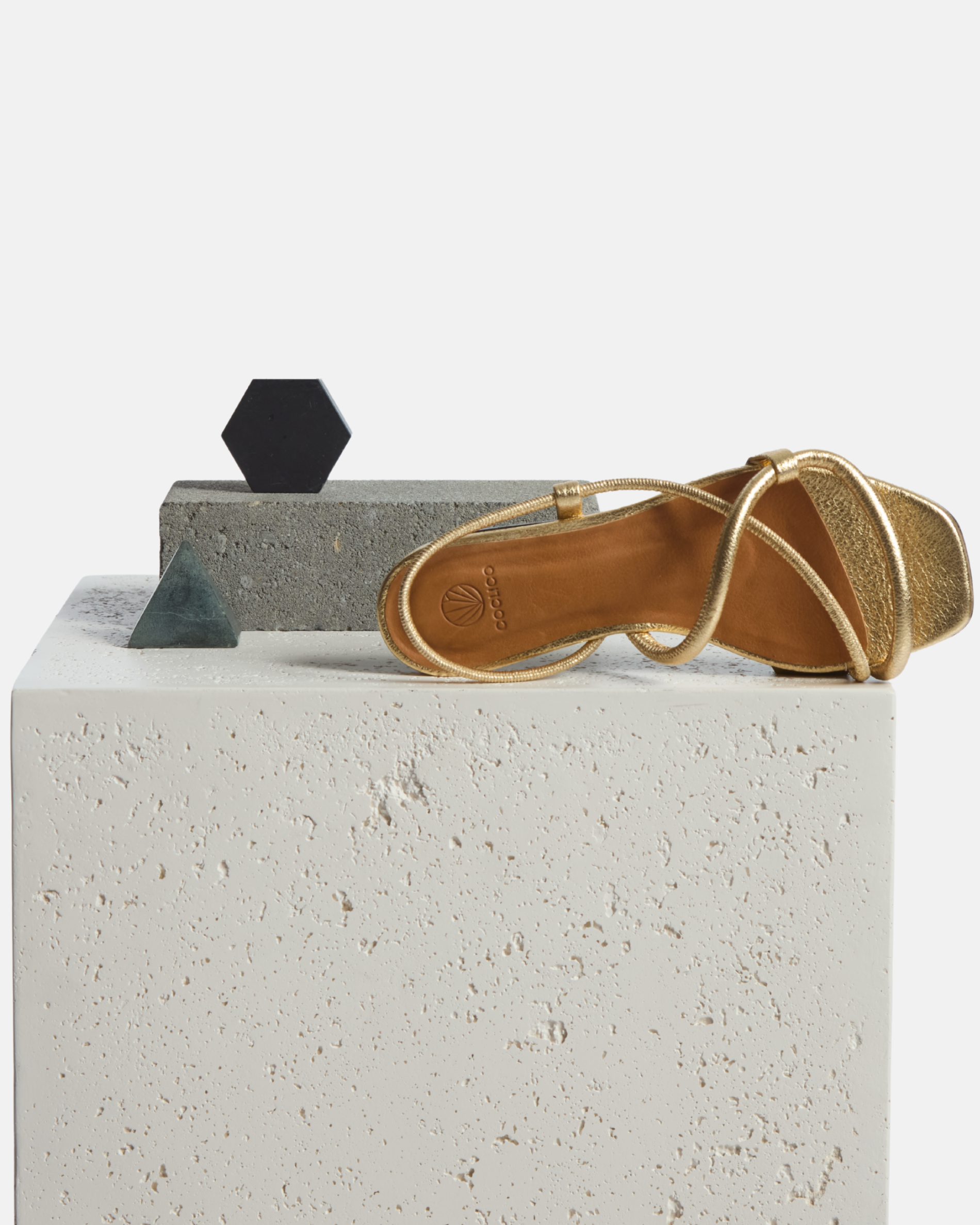 Sandal on a plinth: crinkled metallic gold flat sandal with slim cylindrical padded straps and an elasticated slingback, square toe, on a white background.