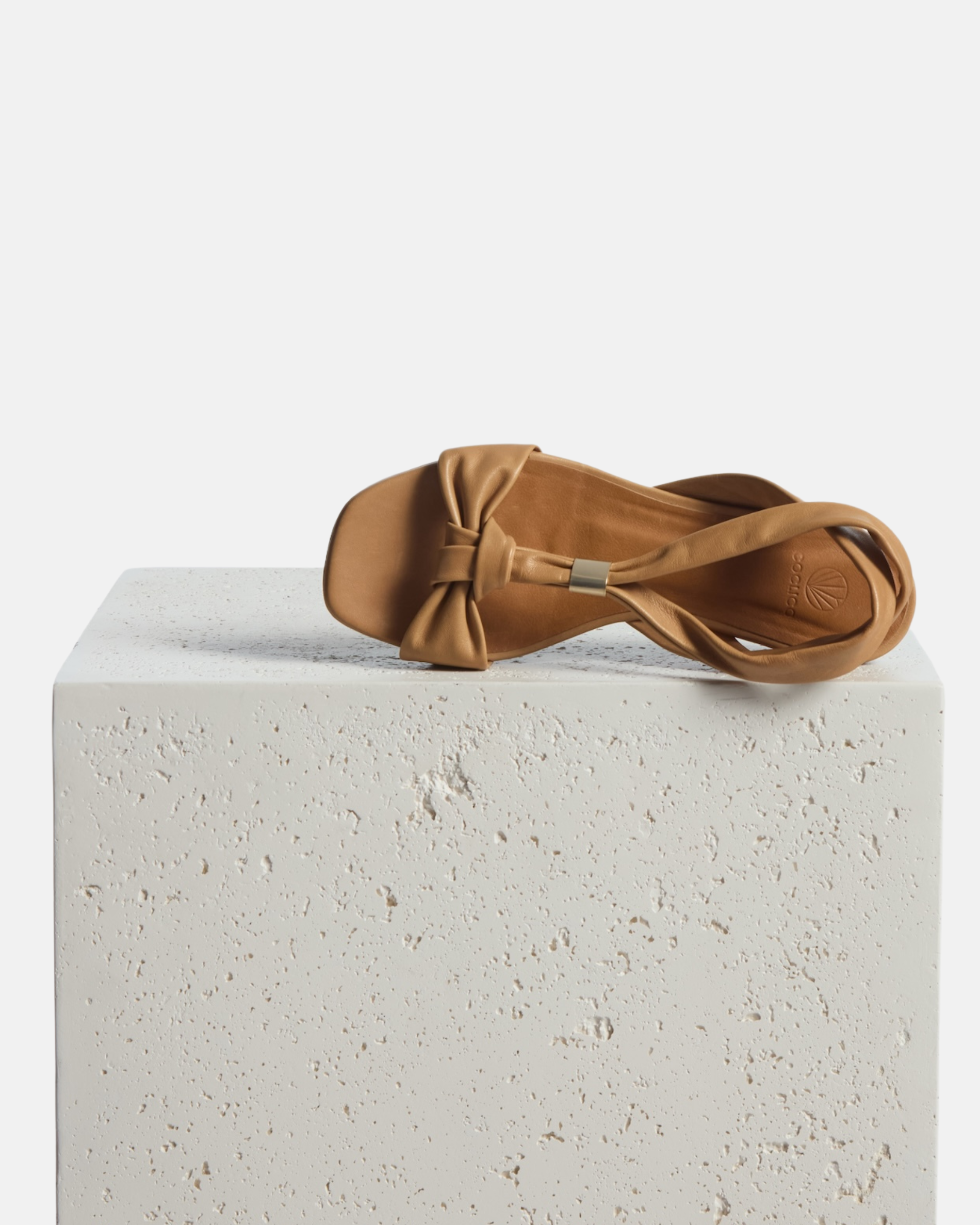 Sandal on plinth: tan leather flat sandal with tubular straps, an adjustable brushed-metal keeper at the center strap, toe knot detail, and a square toe, on a white background.