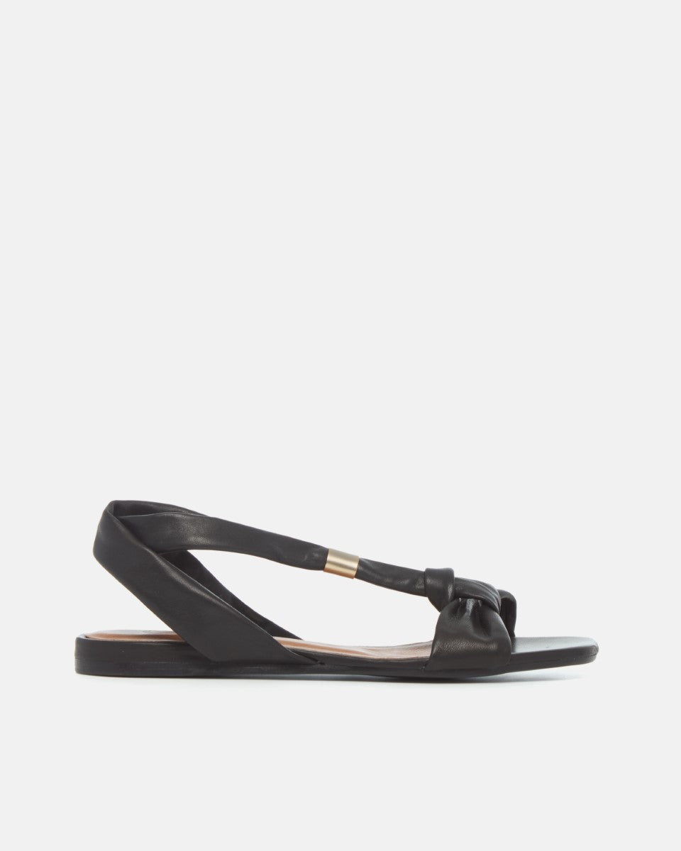 Side view of a black sandal with tubular strap and gold slide at the center of the strap, toe knot detail and a square toe on a white background
