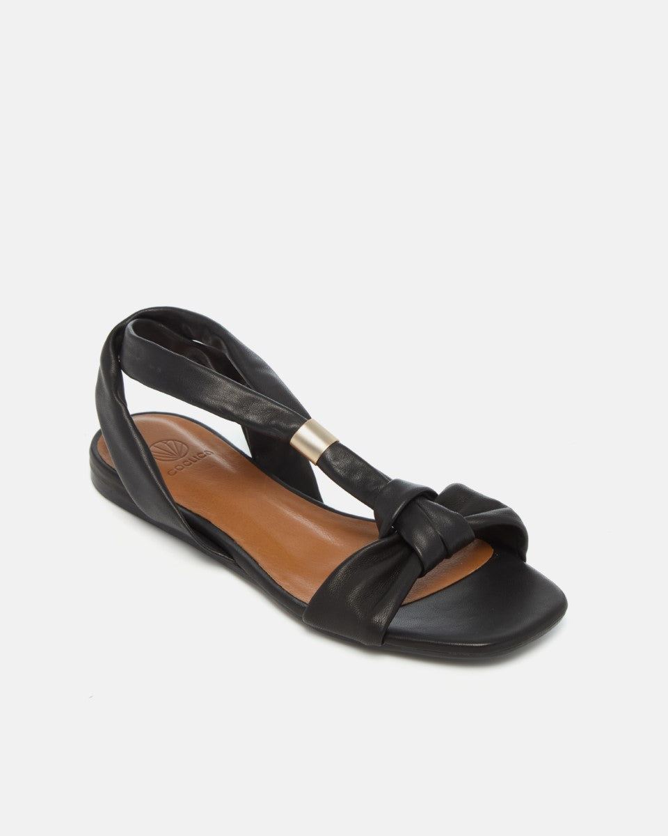 Angled view of a black sandal with tubular strap and gold slide at the center of the strap, toe knot detail and a square toe on a white background