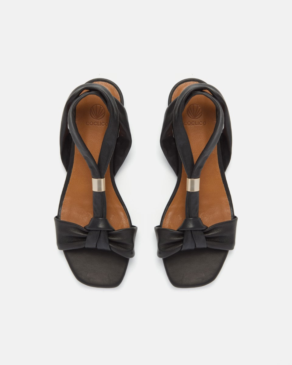 Front view of a pair of black sandals with tubular strap and gold metal slide at the center of the strap, toe knot detail and a square toe on a white background