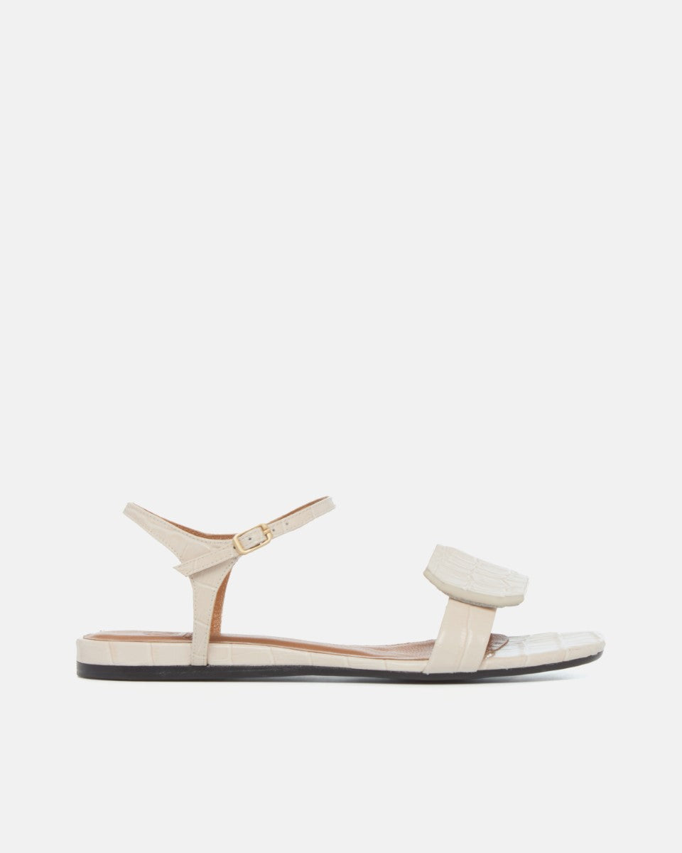 Side view of a bone-colored croc-embossed leather flat sandal with a slim ankle strap and a octagonal accent on the front strap, square toe, on a white background.