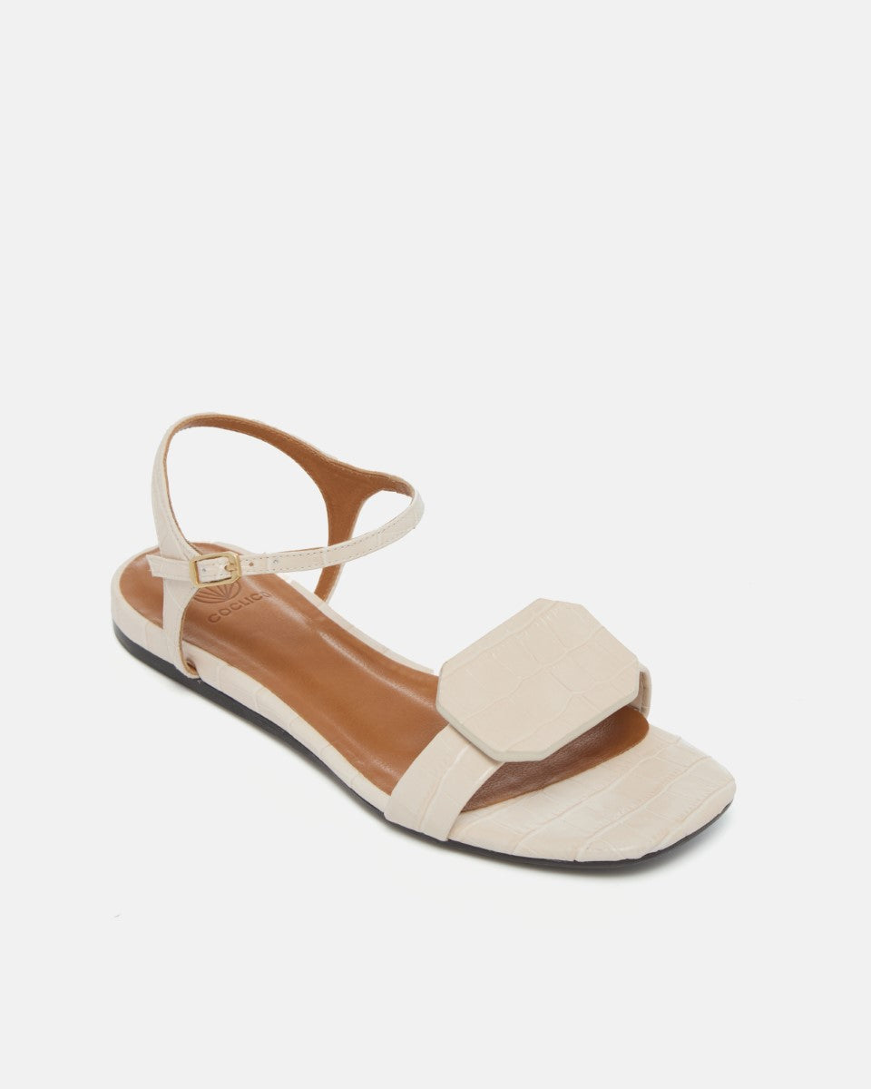 Angled view of a bone-colored croc-embossed leather flat sandal with a slim ankle strap and a octagonal accent on the front strap, square toe, on a white background.
