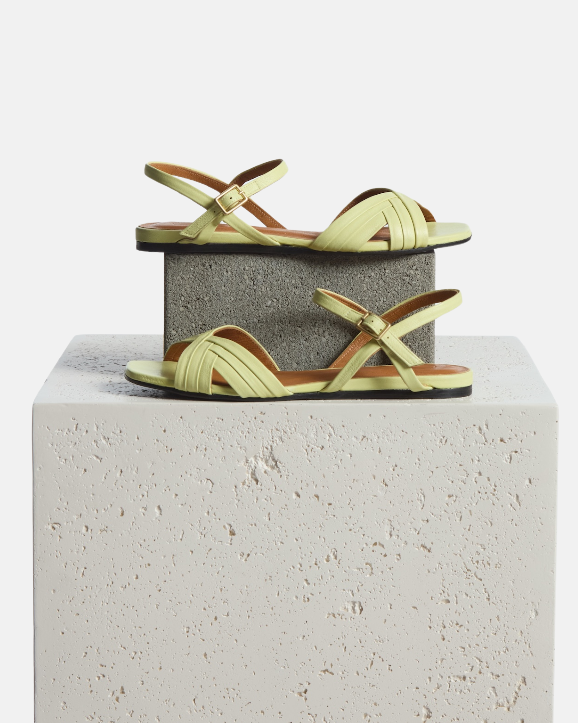 Sandal on plinth: pale yellow leather flat sandal with slim crisscross toe straps, buckle quarter strap, and square toe, on a white background.