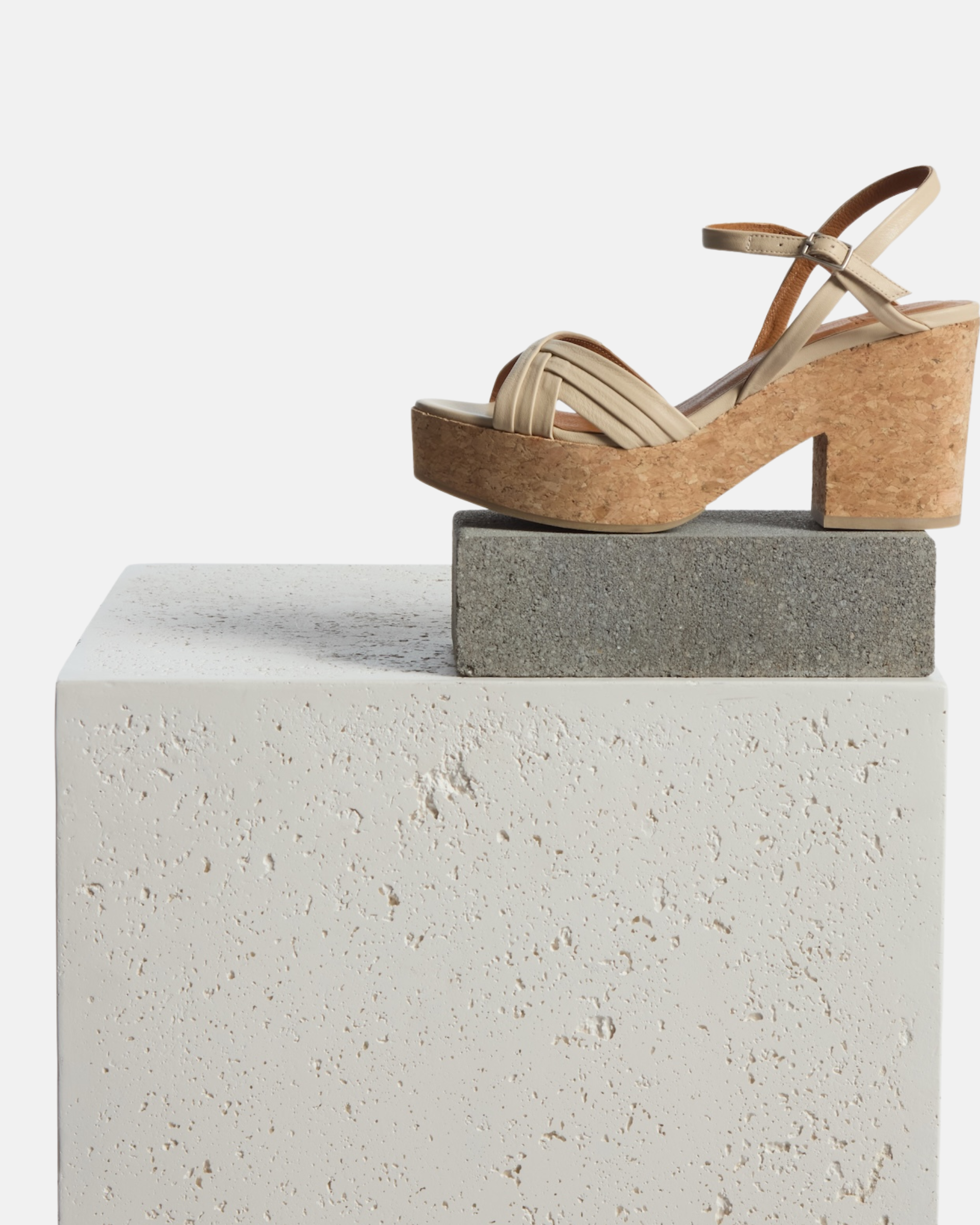 Beige strappy sandals with a cork block heel, featuring a crisscross design on the front and an adjustable ankle strap, resting on a textured white platform.