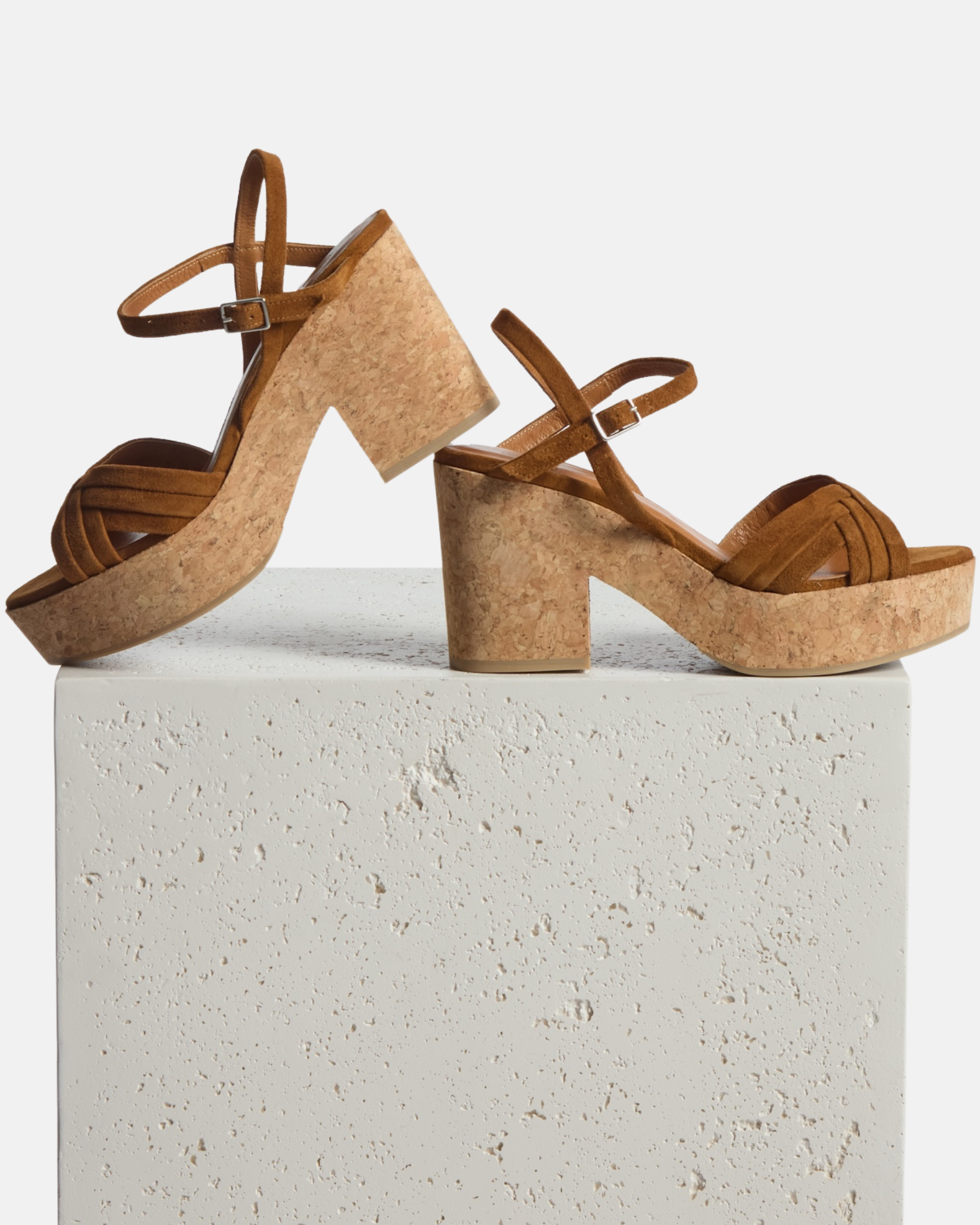 A brown sandal with a cork block heel and platform, featuring a strappy suede upper and an adjustable ankle strap, displayed on a textured white pedestal.