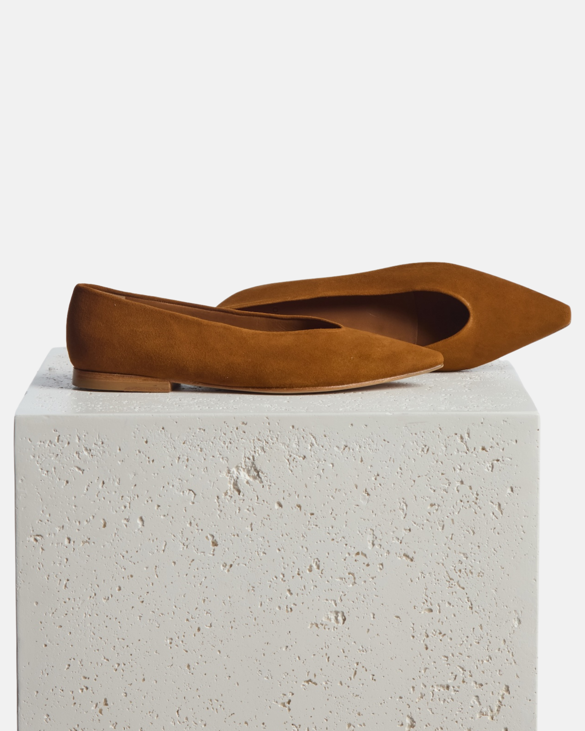 Shoes on a pedestal: warm brown suede pointed-toe flat with clean lines and a flexible leather sole, on a white background.