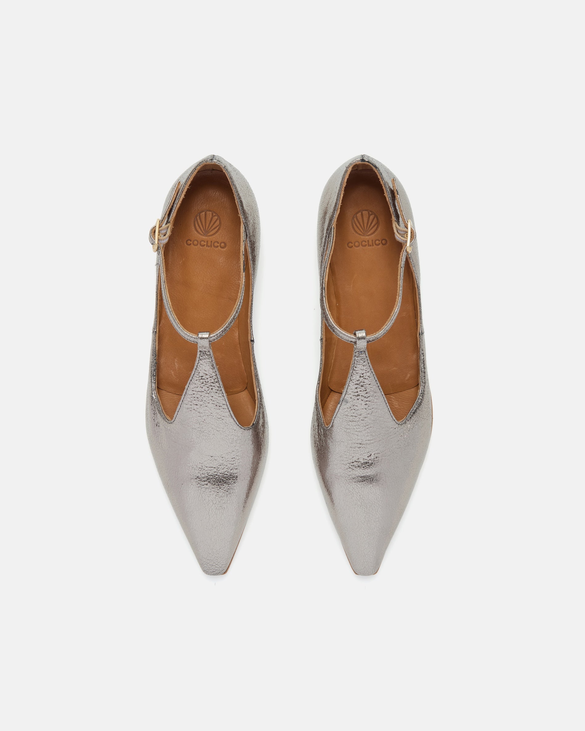 Top view of a pointed-toe silvery metallic T-strap flat with adjustable buckle and leather sole on a white background