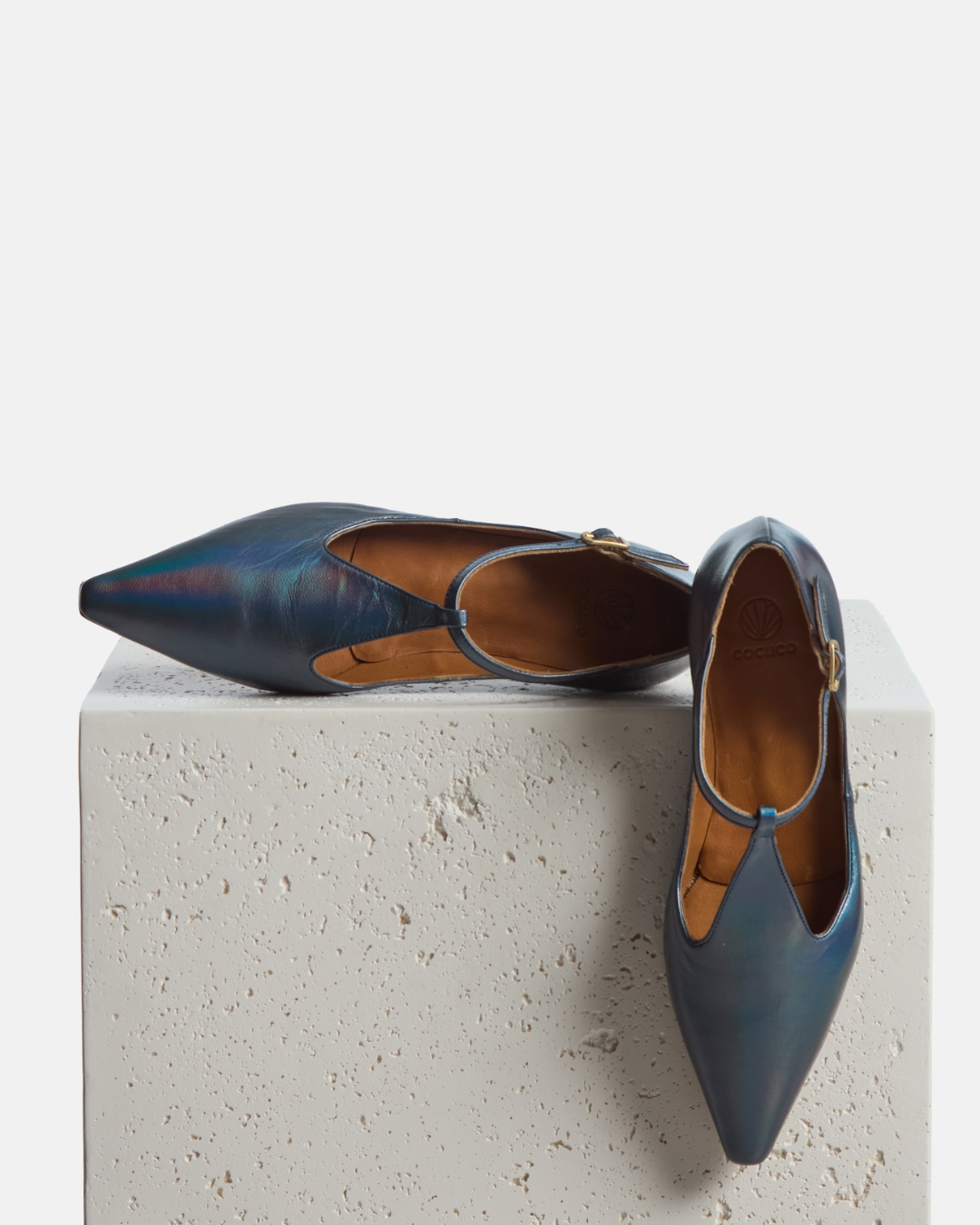 Shoes on a pedestal: pointed-toe iridescent blue metallic T-strap flat with adjustable buckle and leather sole on a white background