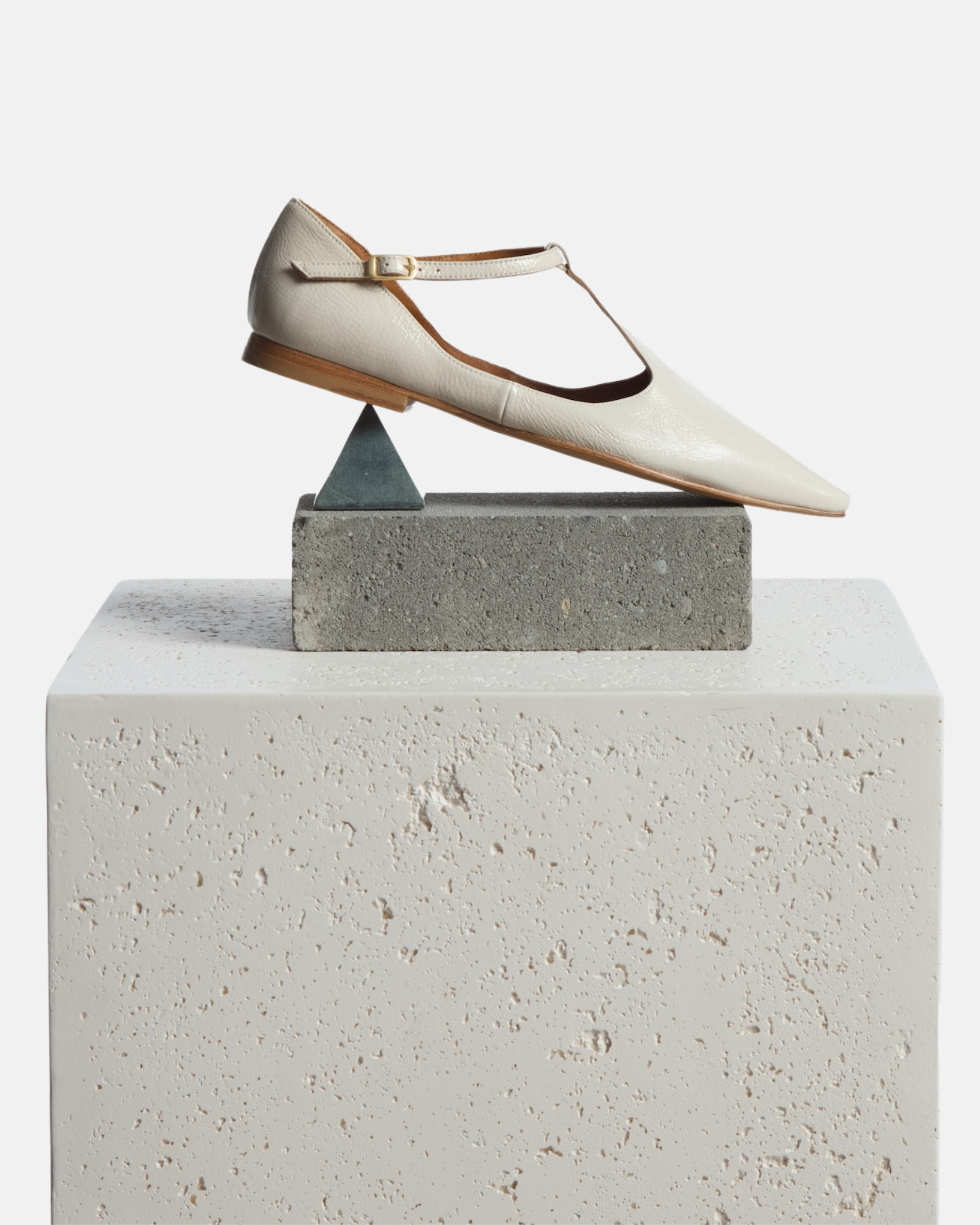 Shoe on a pedestal: pointed-toe off-white patent leather T-strap flat with adjustable buckle and leather sole on a white background