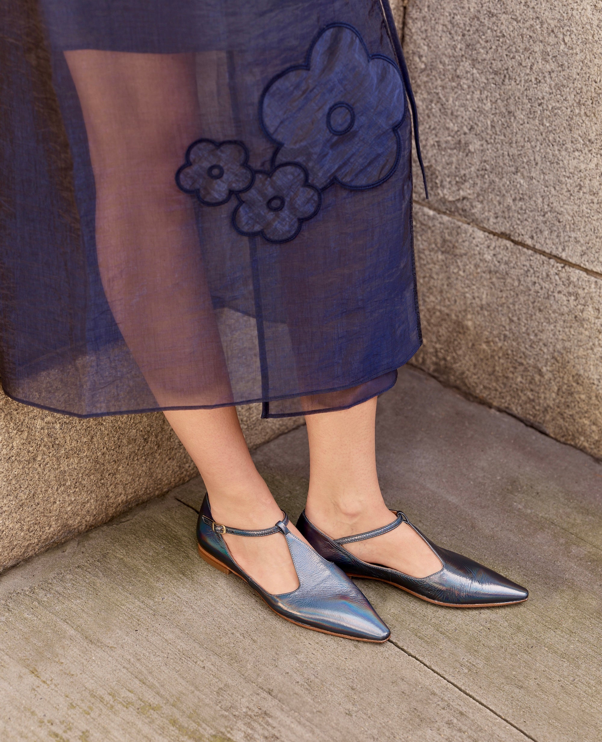 Person wearing a blue floral dress and iridescent flats on a stone pavement.
