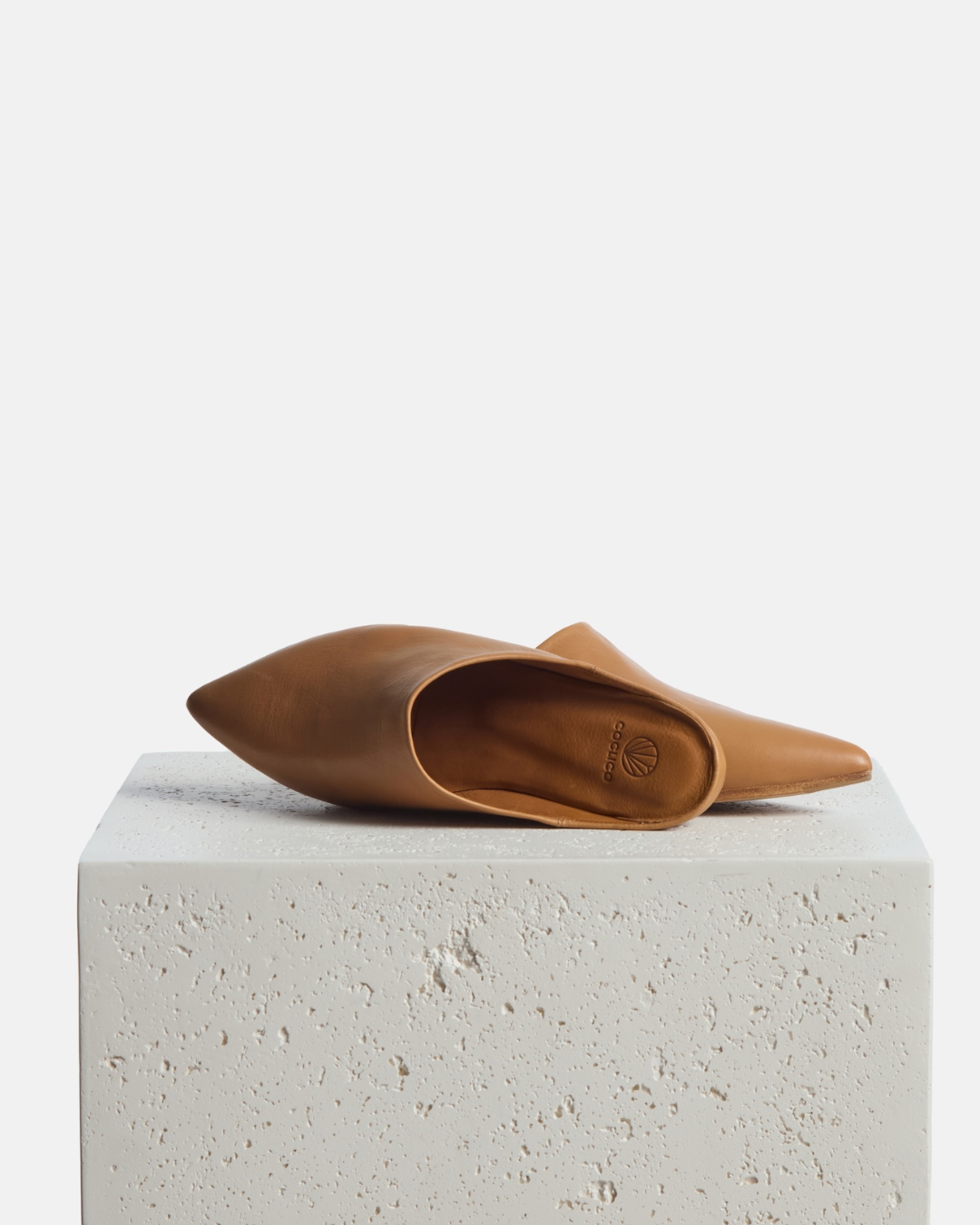 Mules on a plinth: light brown pointed-toe leather mule with a slight built-in wedge and clean minimalist silhouette on a white background