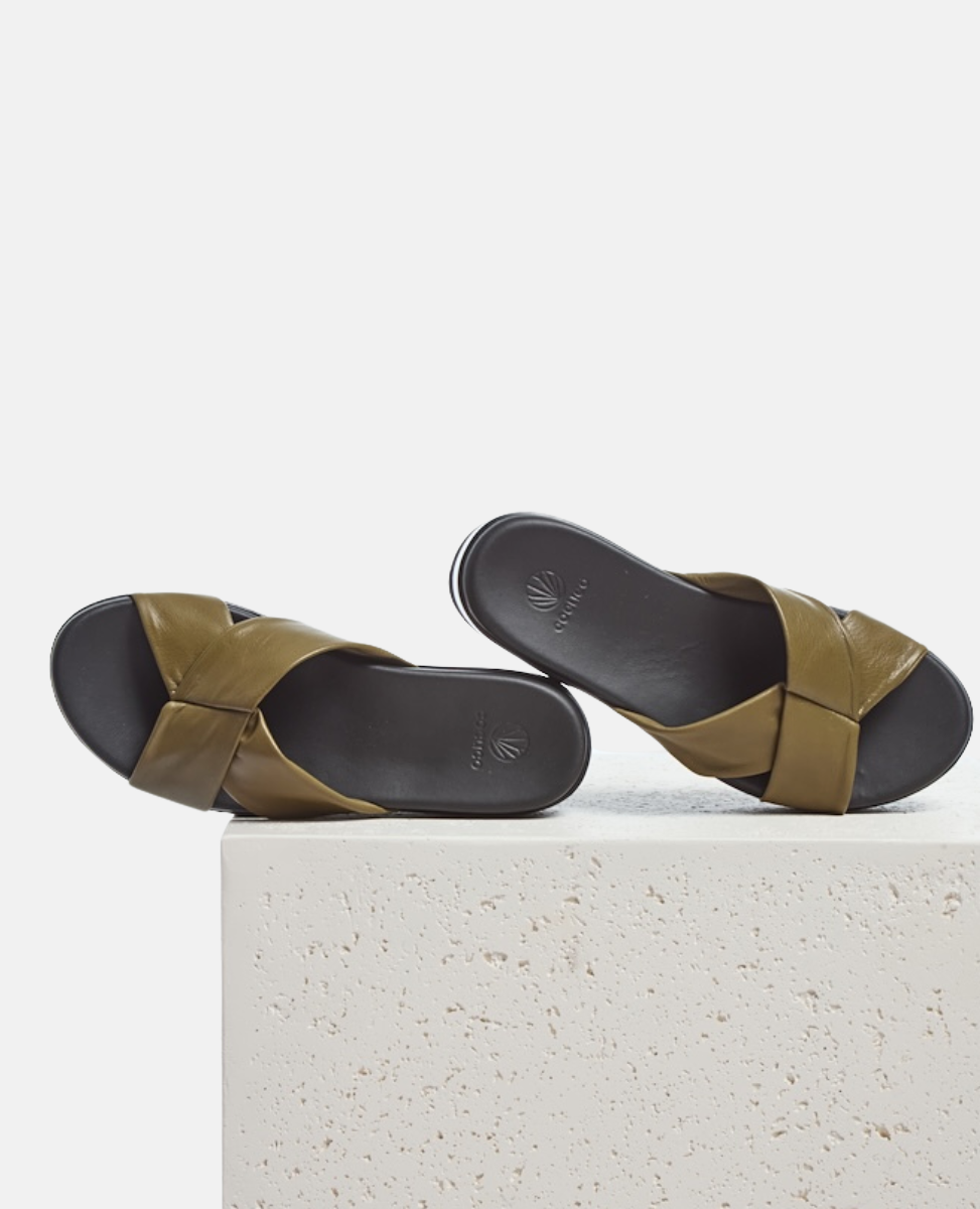 A pair of olive green leather sandals with crisscross straps, a smooth black footbed, and a minimalist design, resting on a textured white surface.
