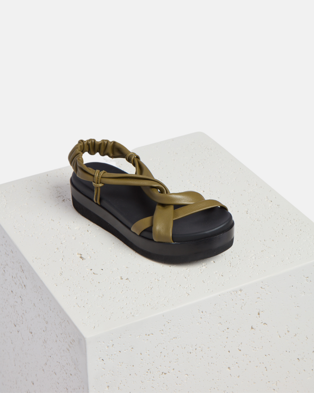 Shoe on a plinth: A sandal featuring a thick black platform sole, olive green leather straps with a crisscross design, and an elasticized back strap for a secure fit.