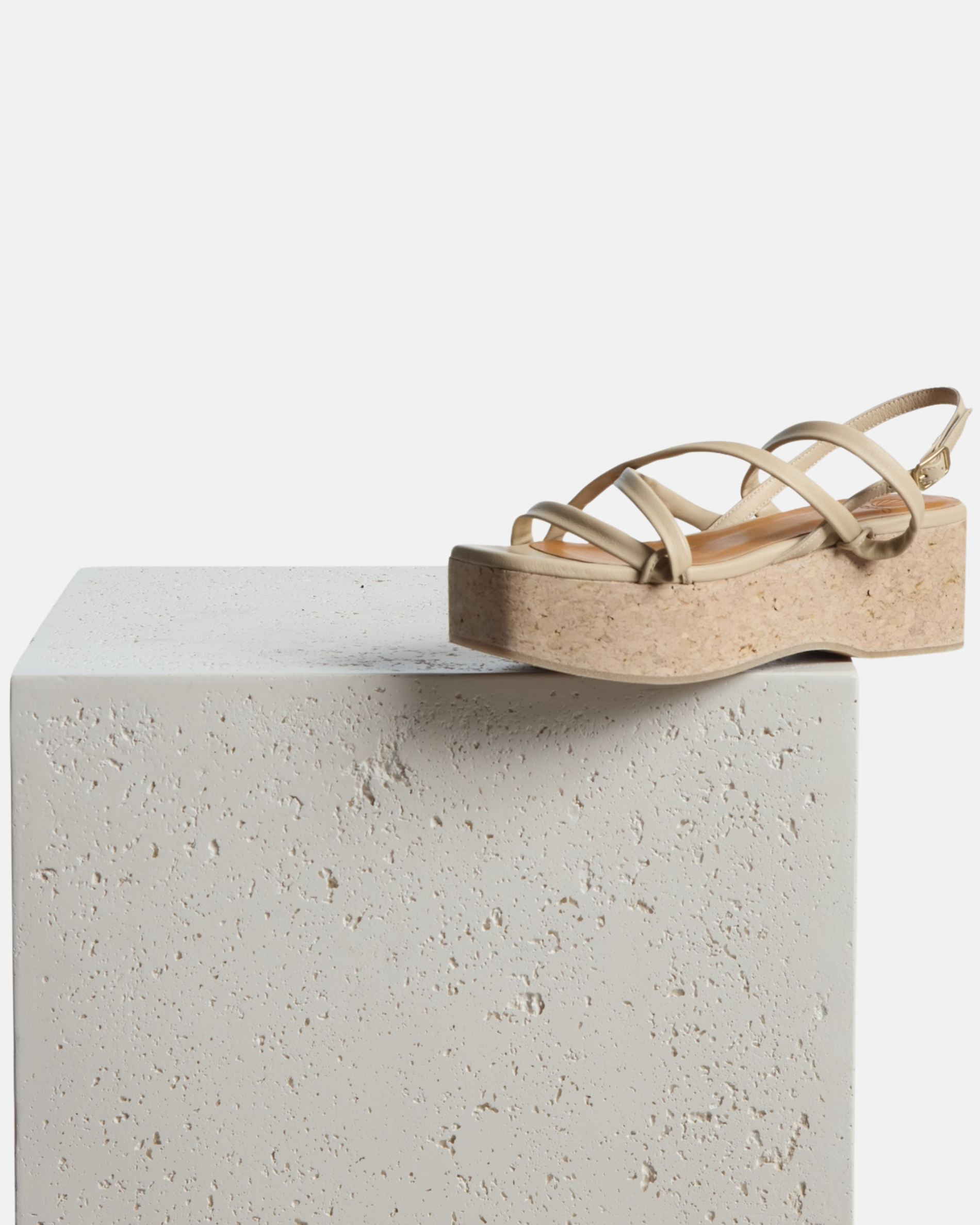 Cork wedge sandal perched on the side of a pedestal: off-white leather cork wedge sandal with slim crisscross straps, adjustable slingback, and a sculptural cork platform sole