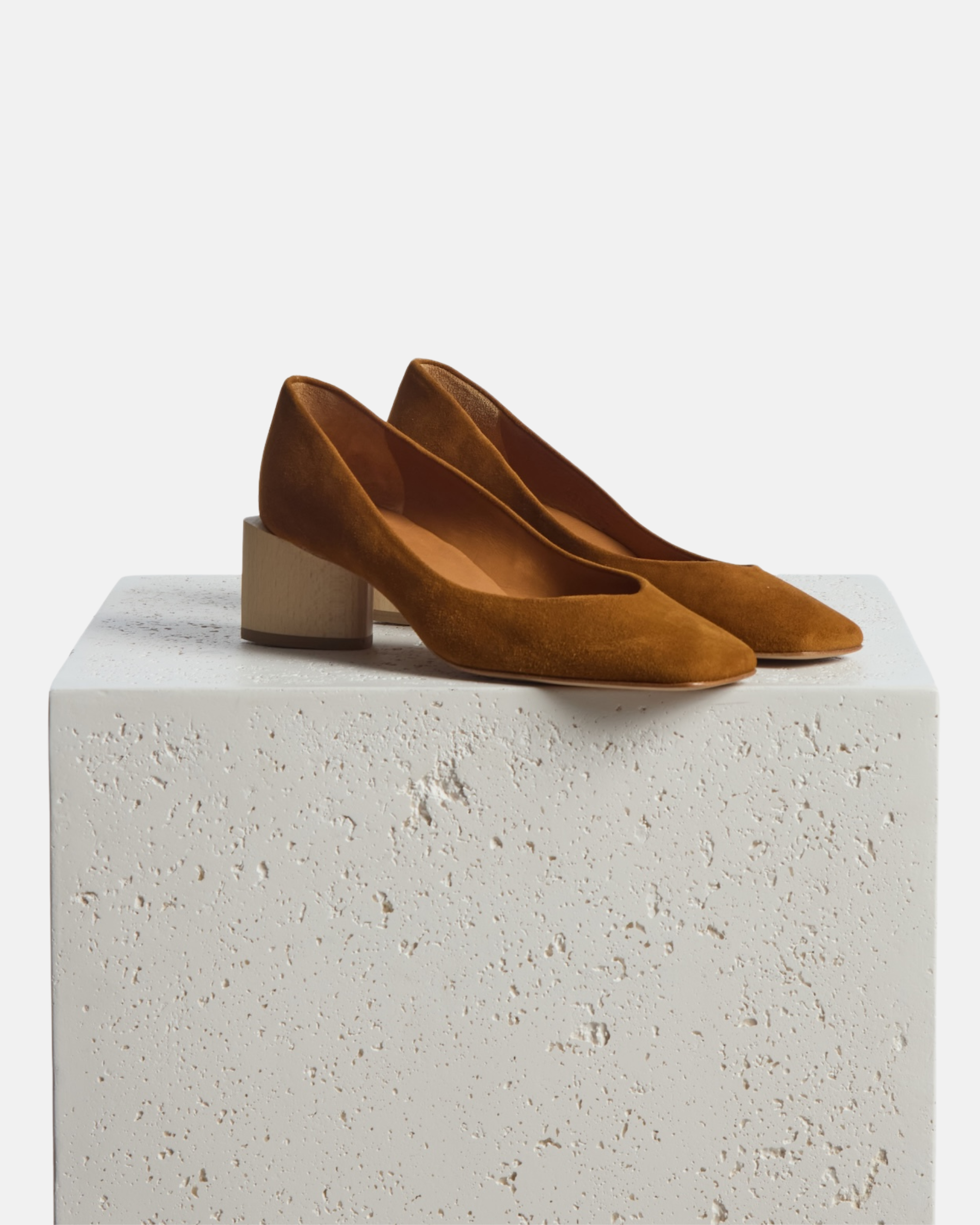 Shoes on the edge of a plinth: a brown suede pump with a square toe and a solid architectural wood block heel on a white background