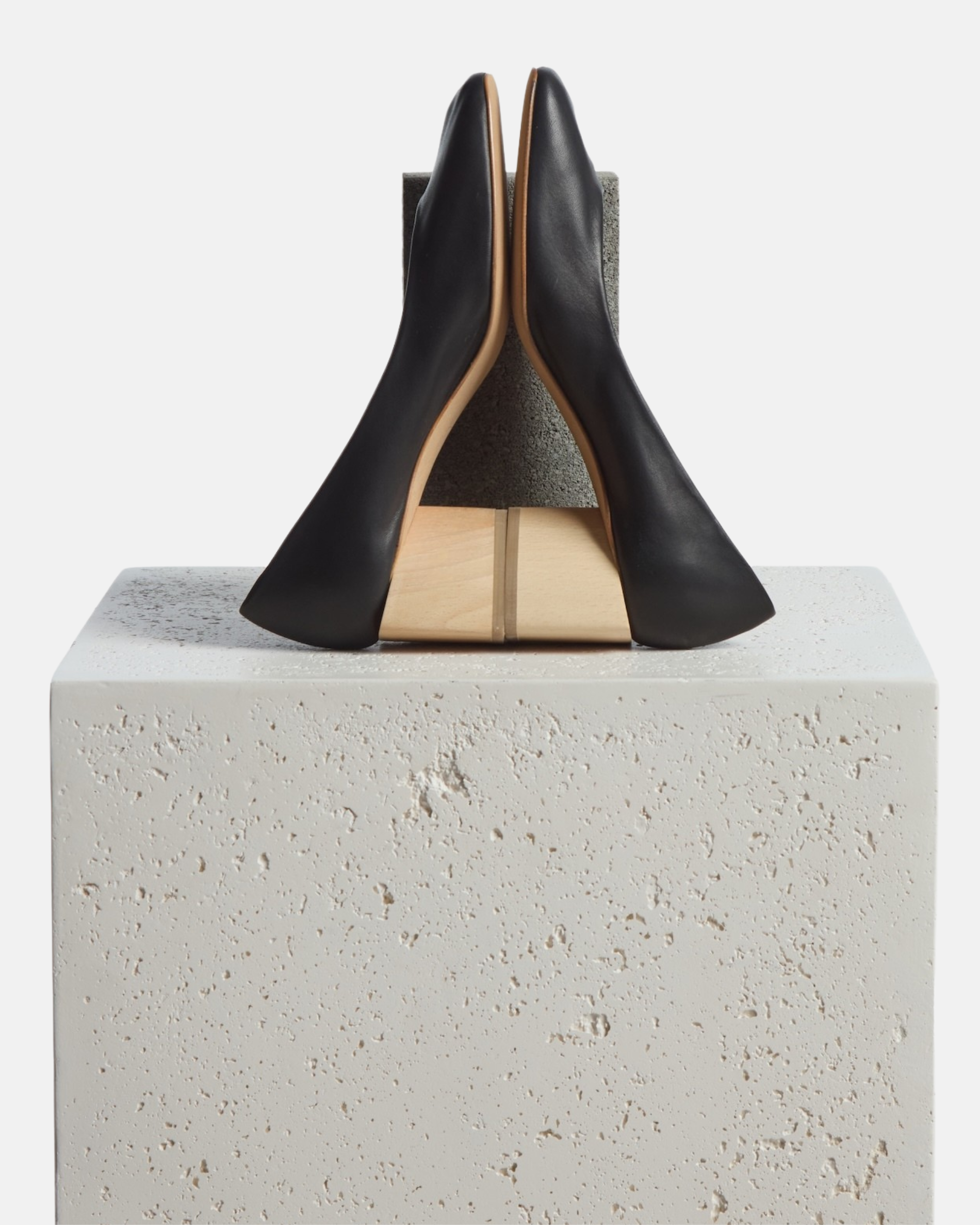 Shoes arranged creativly on a plinth: black leather pump with a square toe and a solid architectural, natural wood block heel on a white background