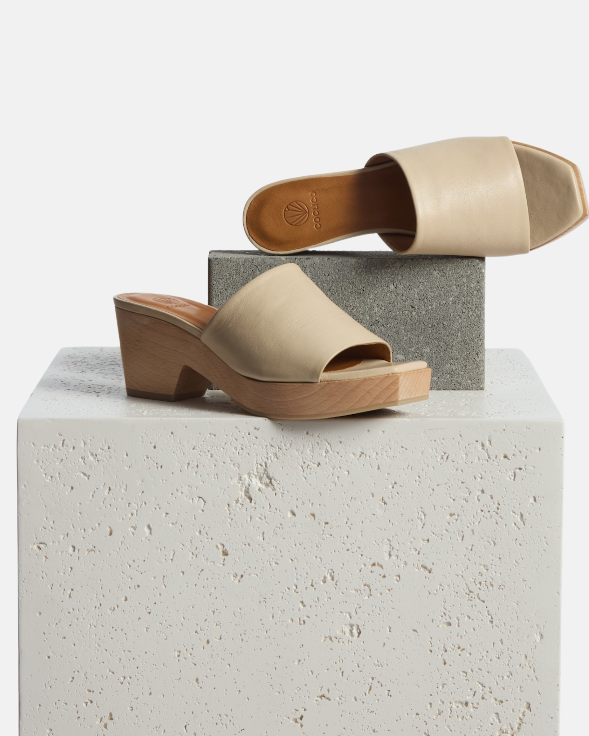 Clogs on a plinth: A pair of off-white leather, open-toe slide sandal clogs, featuring a chunky wooden block heel and a minimalist design.