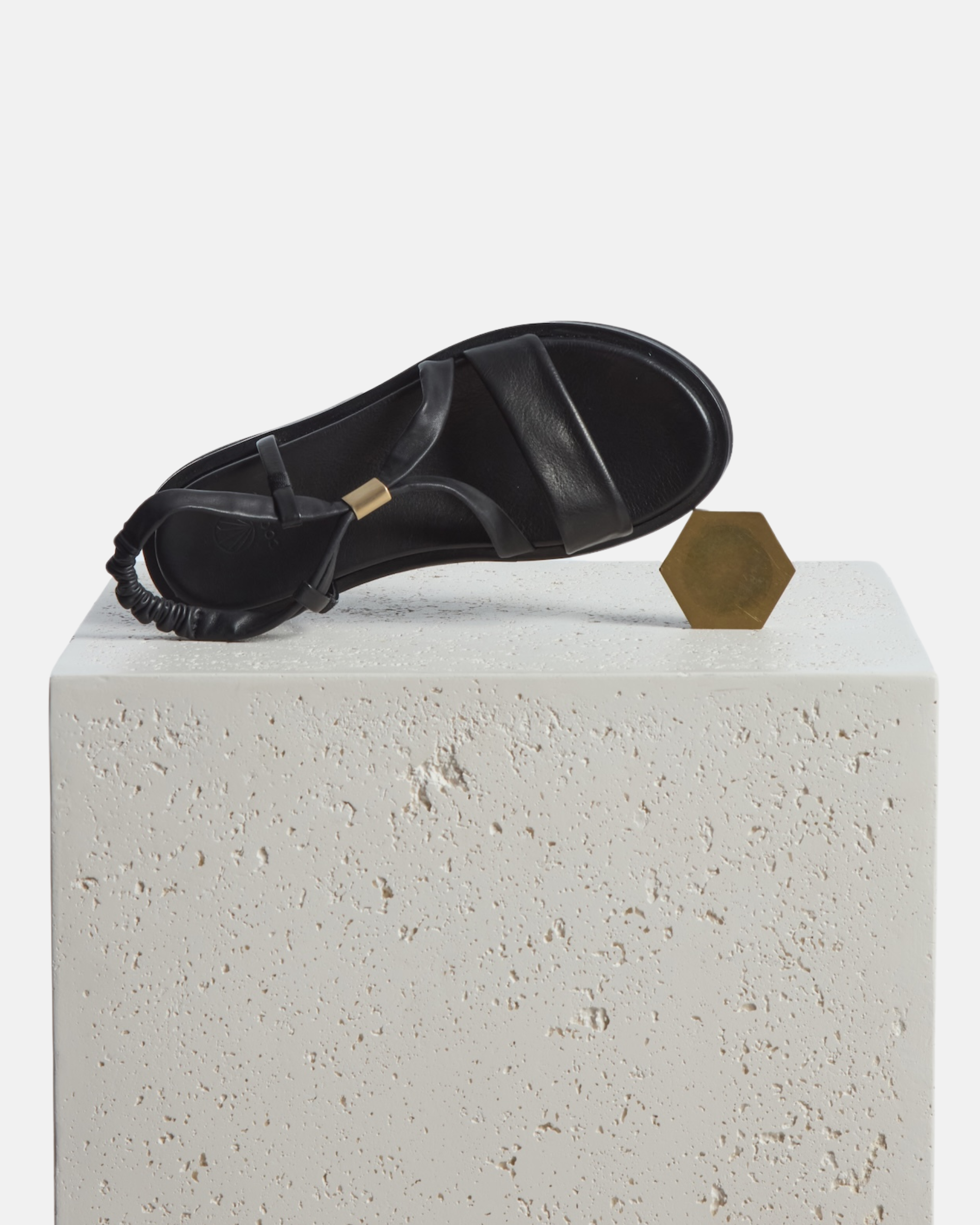 Footbed shoe on a plinth: black leather slingback sandal with soft tubular straps, brushed-metal keeper detail, and a cushioned platform sole on a white background