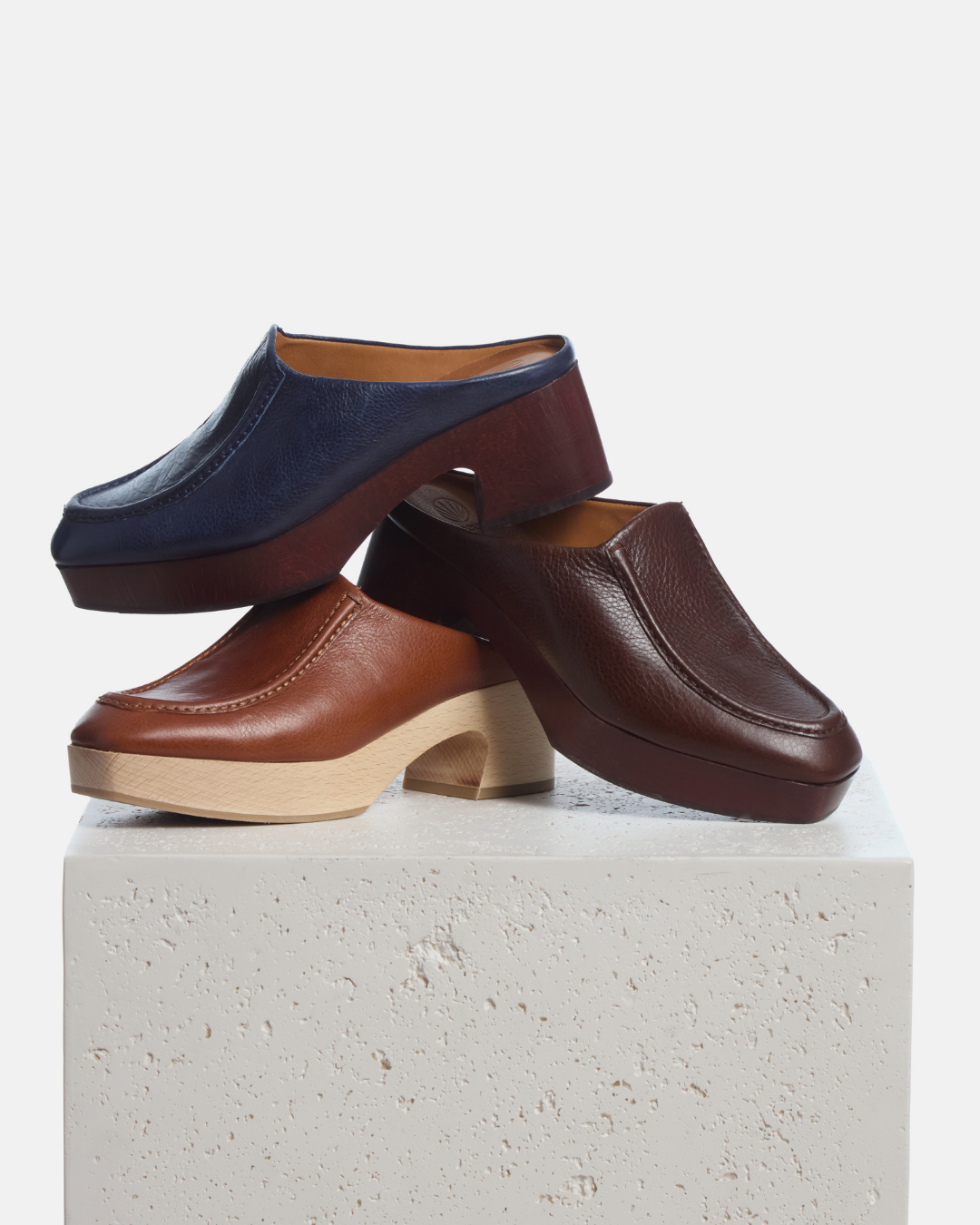 Three leather clog-style mule shoes stacked on a white stone pedestal against a light gray studio background. The shoes include navy blue, tan-brown, and dark brown options with stacked platform heels and light beige soles.