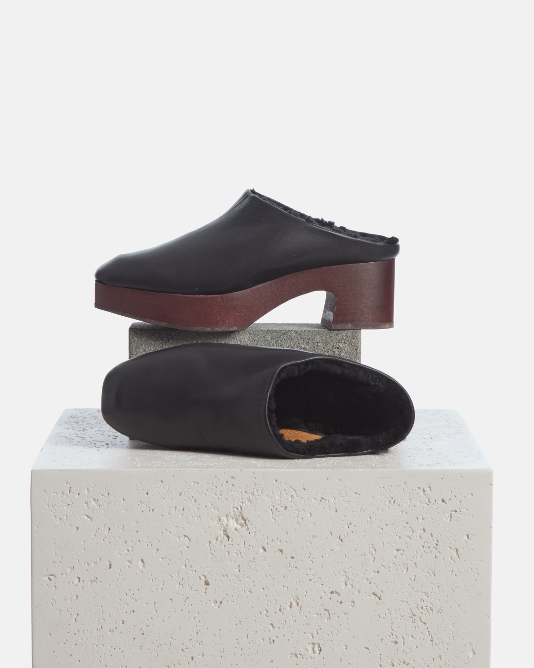 Coclico Quince Shearling Clog in black leather, shearling lined clog mules styled on a stone plinth