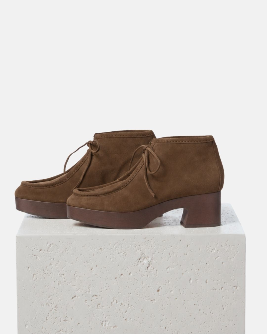 A pair of brown suede lace-up clogs with dark brown wood platform heels, displayed on a textured white stone block against a clean white background.