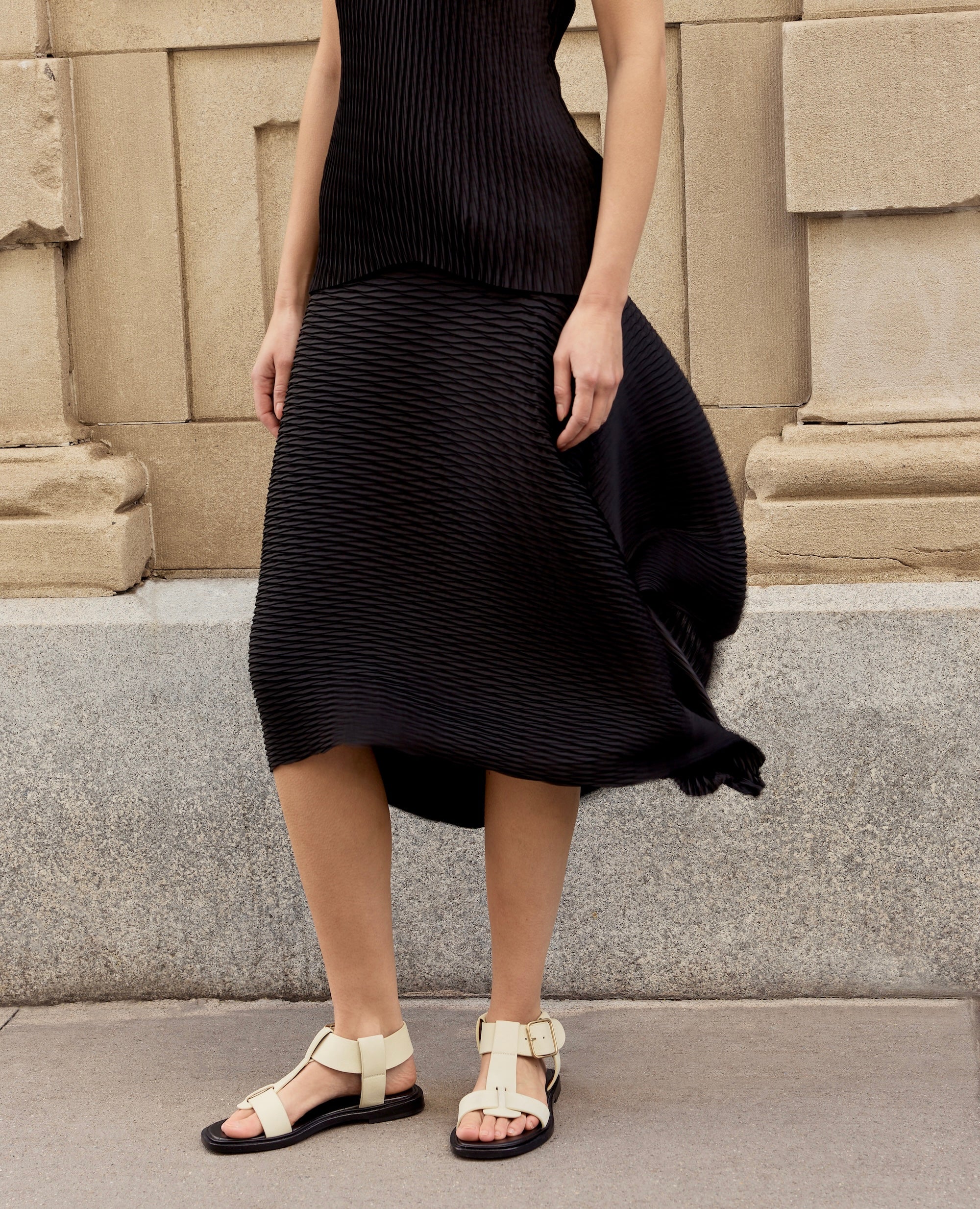 Person wearing a black dress nd white t-strap flat sandals standing against a stone wall.
