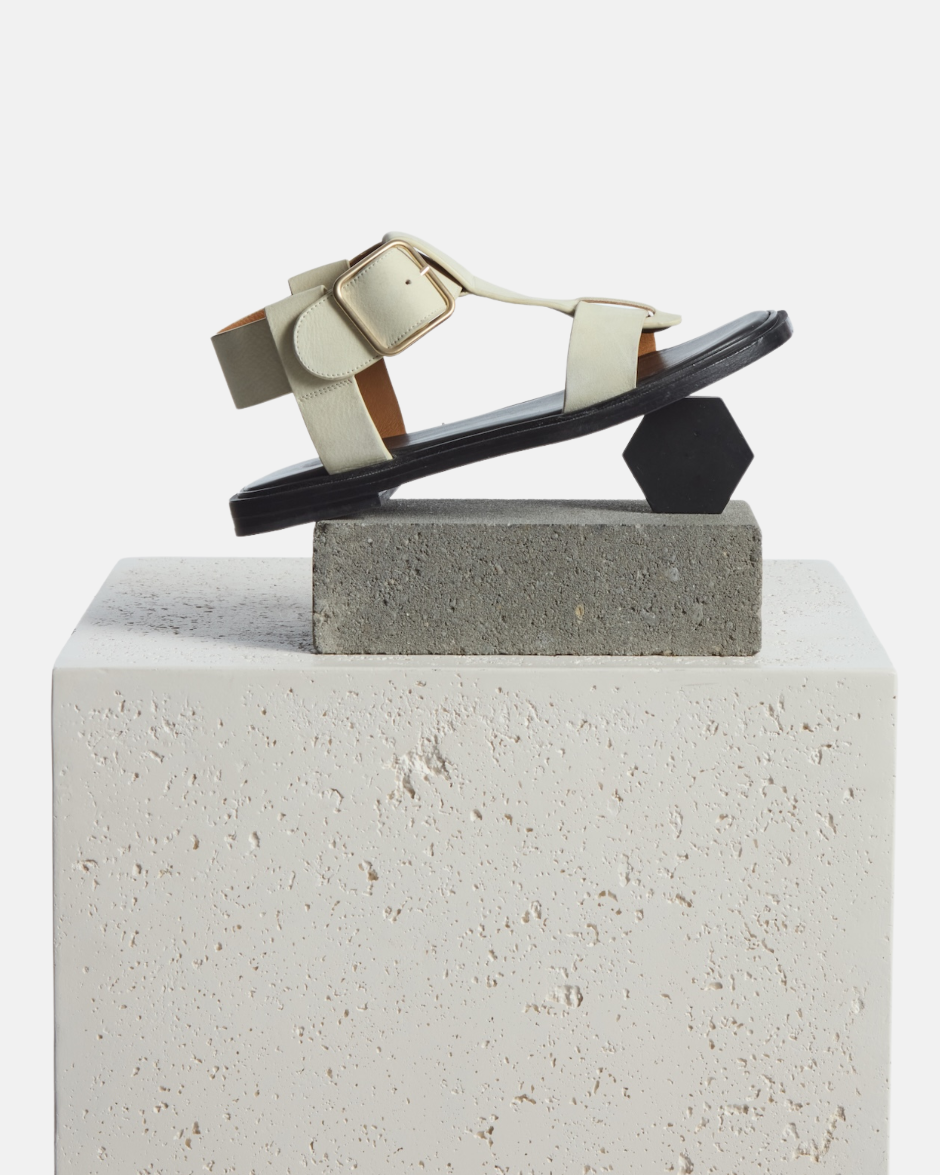 shoe on plinth: white nubuck leather T-strap sandals with wide straps, oversized buckle, cushioned leather footbed, and substantial sole.