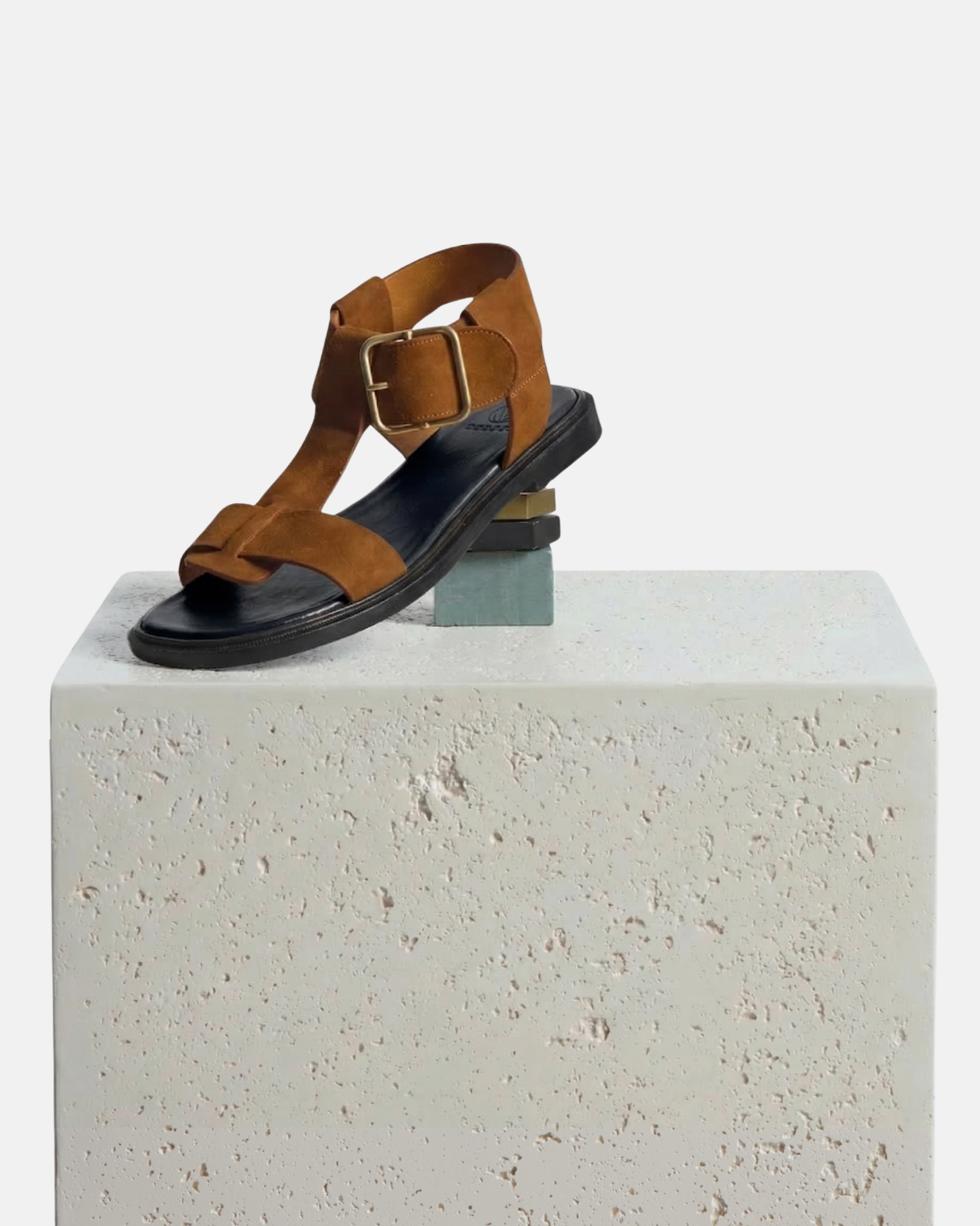 Shoe arranged on a plinth: brown suede T-strap sandal with wide straps, oversized buckle, cushioned leather footbed, and substantial sole