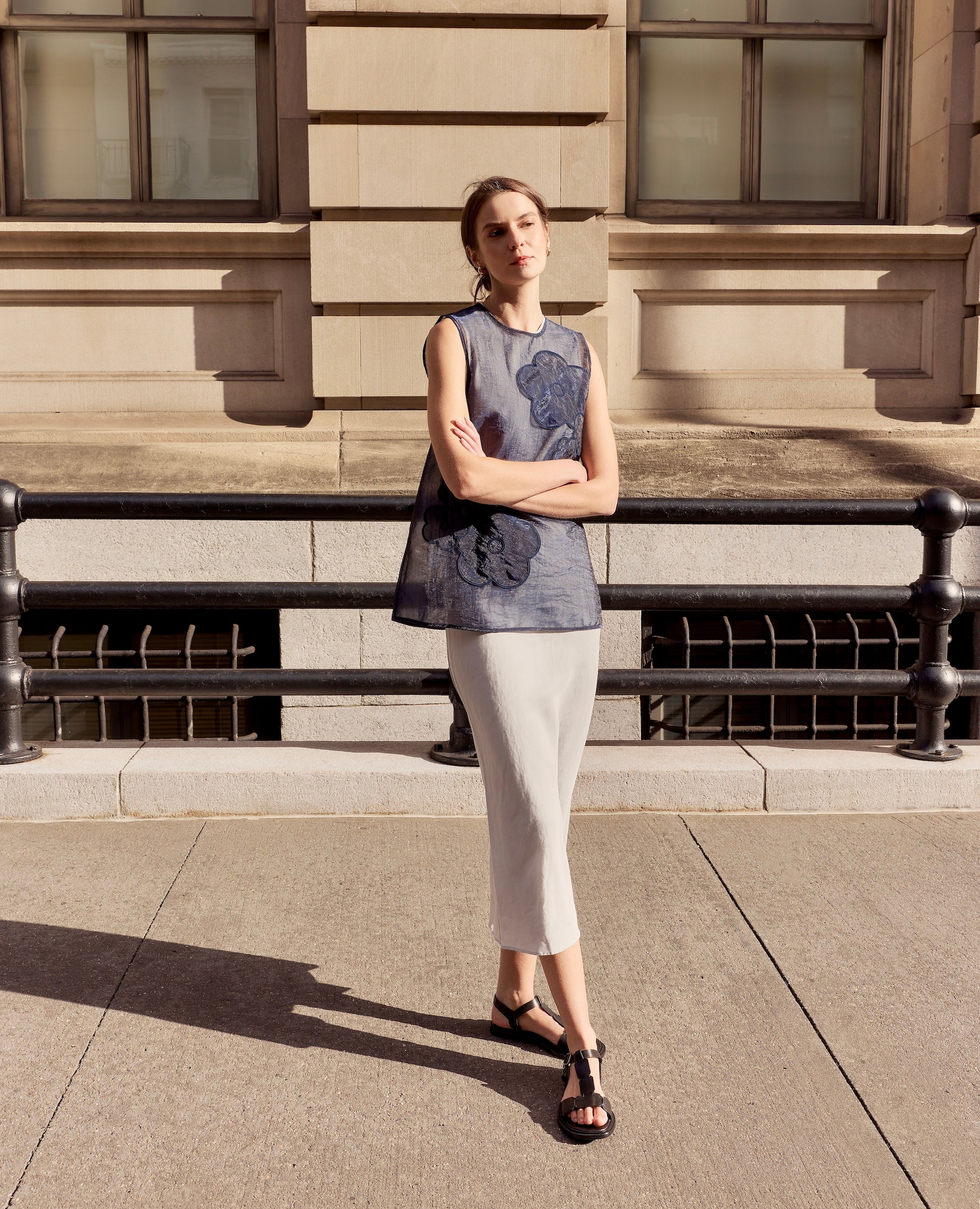 Woman standing in front of a building with a floral sleeveless top, black t-strap sandals, and white pants.