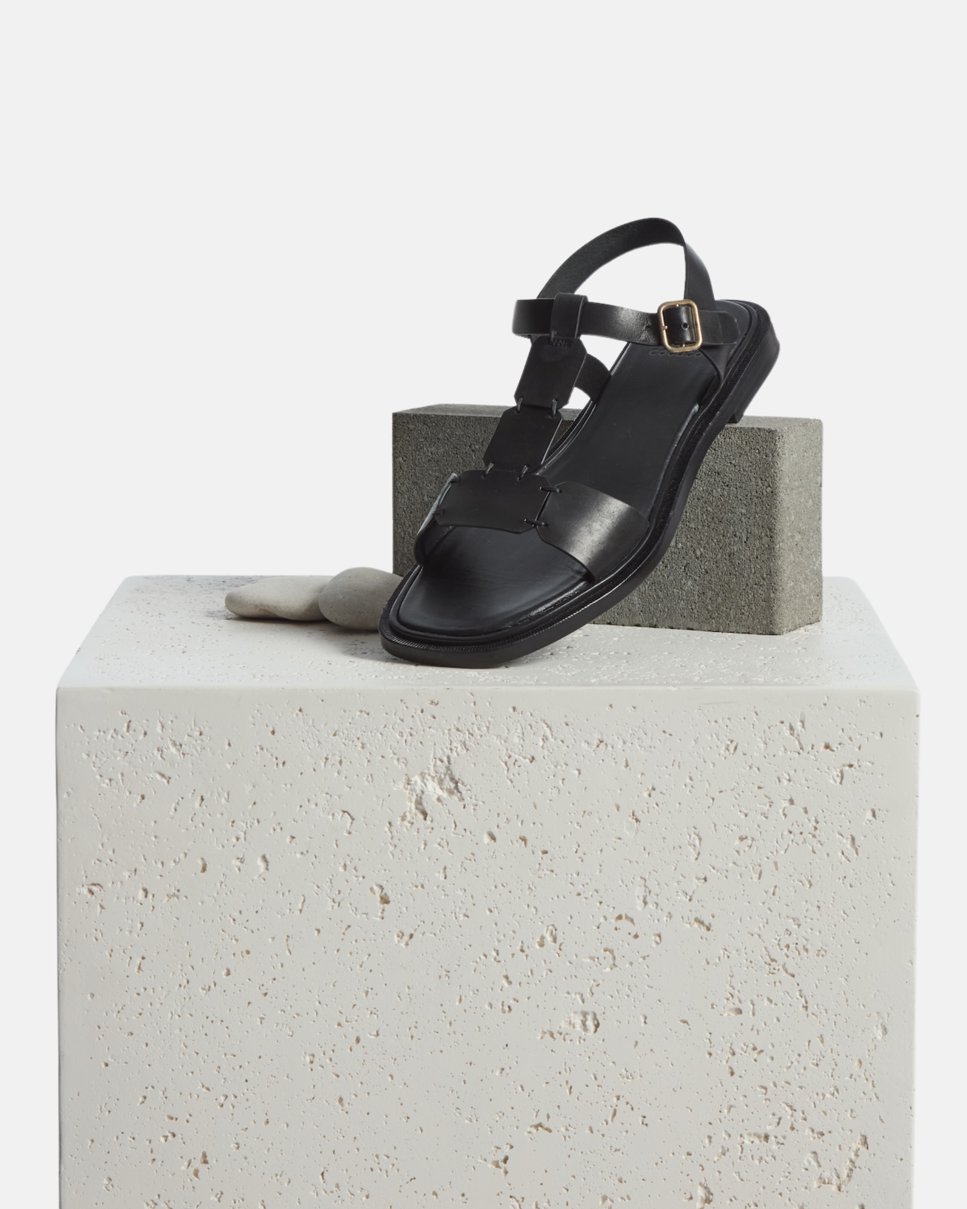 Shoe on plinth: a black leather T-strap sandal with hand-linked panels, slim ankle strap, and a sleek flexible leather sole with a textured welt.
