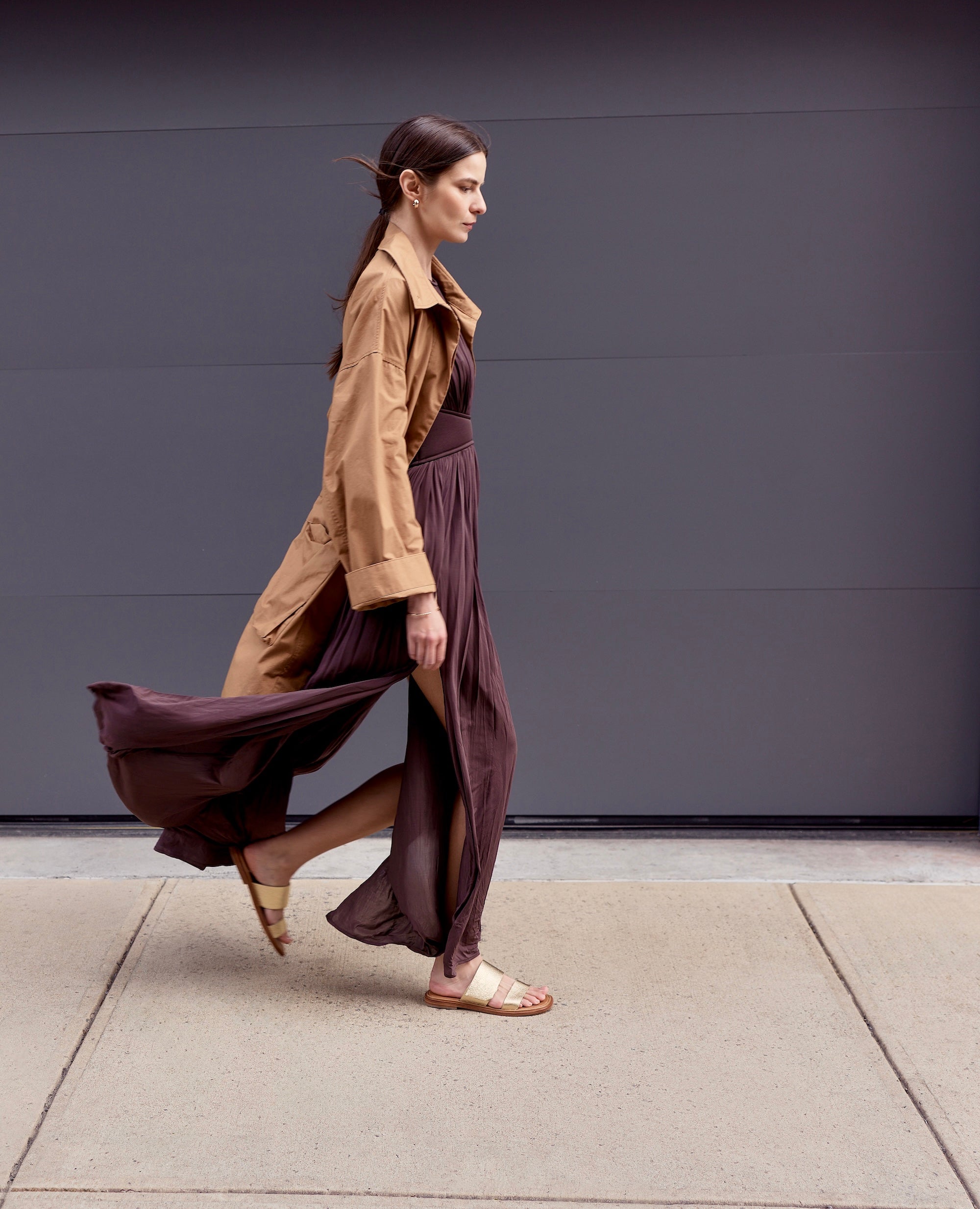 Woman walking on a sidewalk wearing a brown trench coat and long burgundy dress and flat gold sandals.