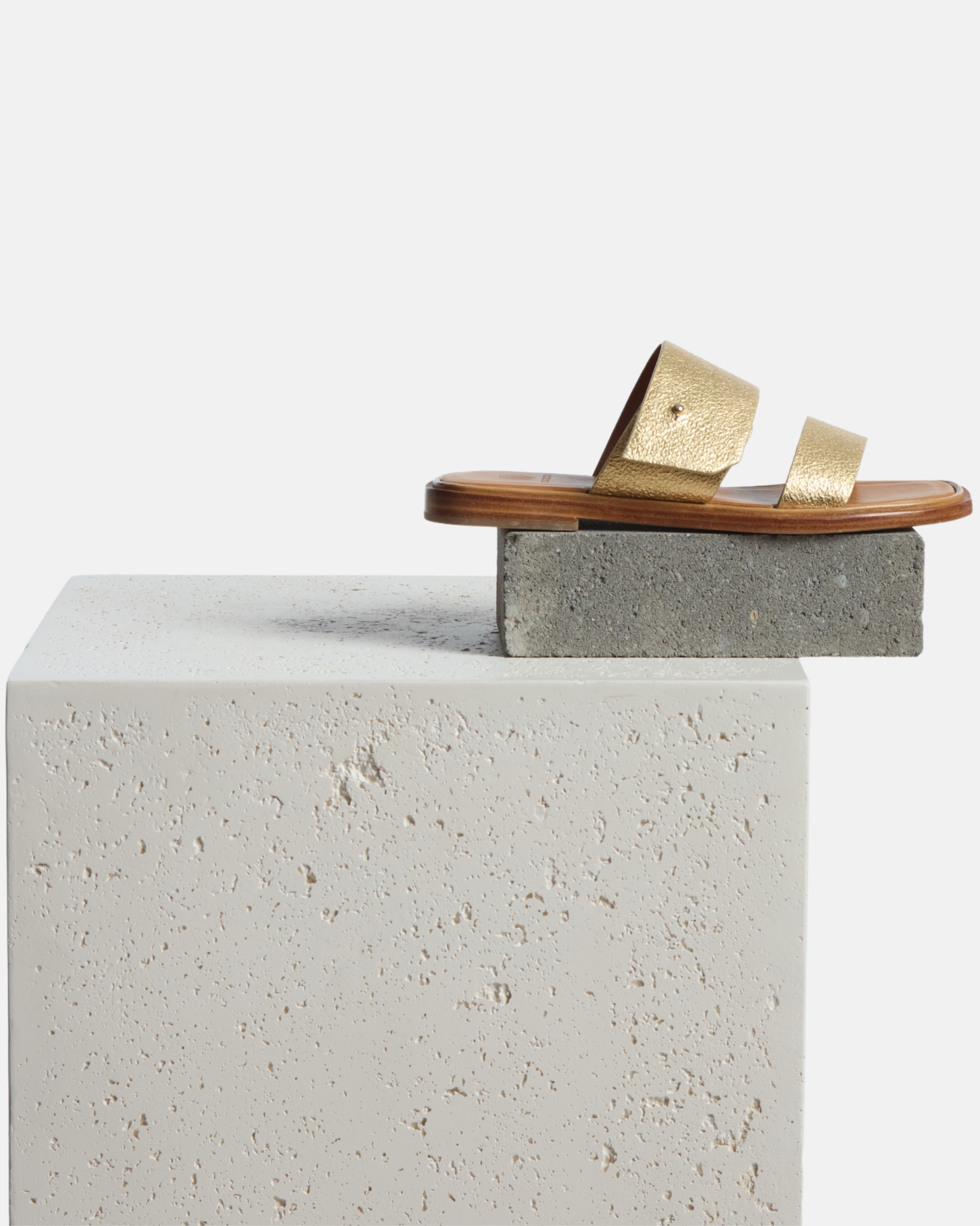 Sandal on a plinth: gold metallic leather slide sandal with two wide straps, a subtle metal peg detail, cushioned leather footbed, and flat leather sole