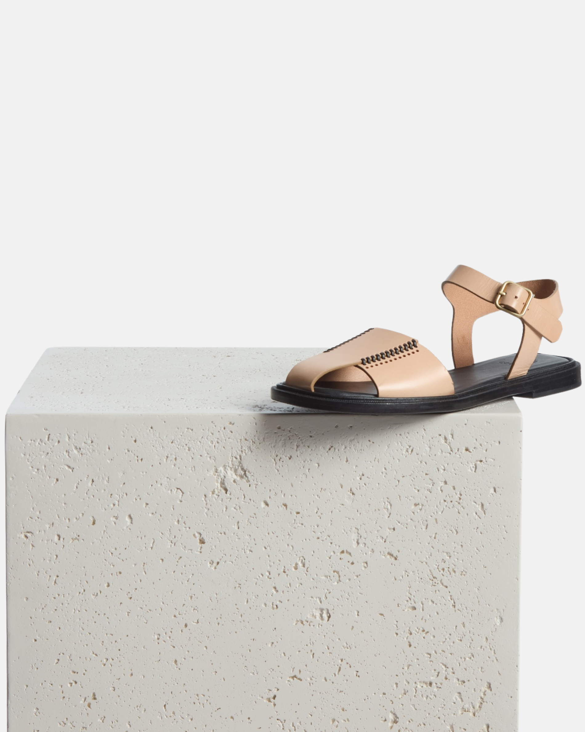 Shoe on plinth:natural leather fisherman sandal with stitched banded straps, slim ankle strap, and a black flexible leather sole with textured welt on a white background.