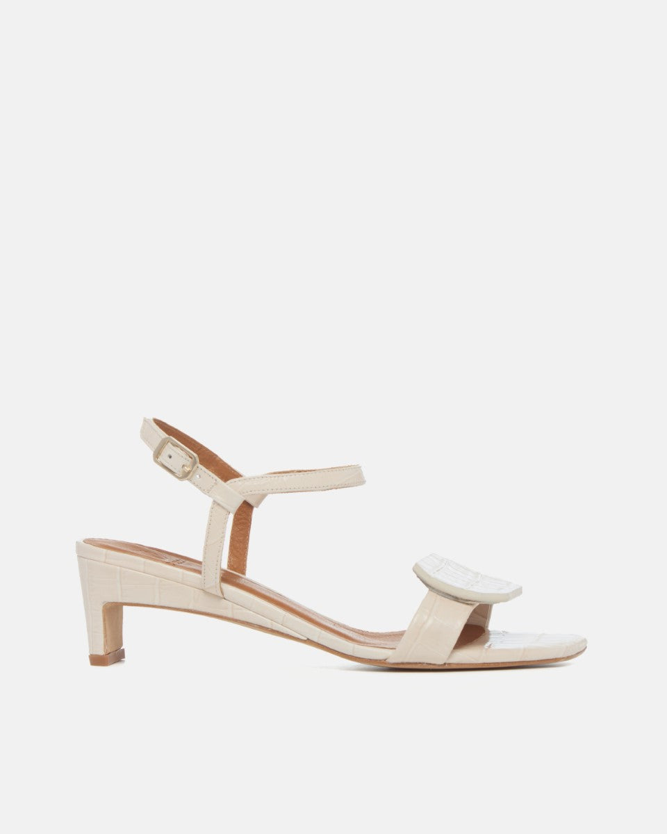 Side view of a bone white croc-embossed leather sandal with a slim ankle strap, octagonal toe detail, and a low wrapped demi heel on a white background.