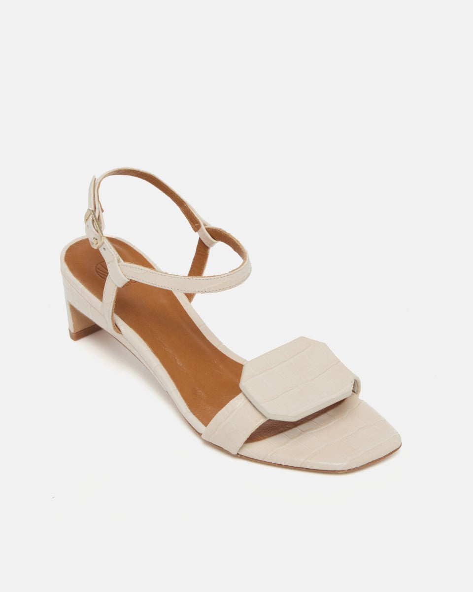 Angled view of a bone white croc-embossed leather sandal with a slim ankle strap, octagonal toe detail, and a low wrapped demi heel on a white background.