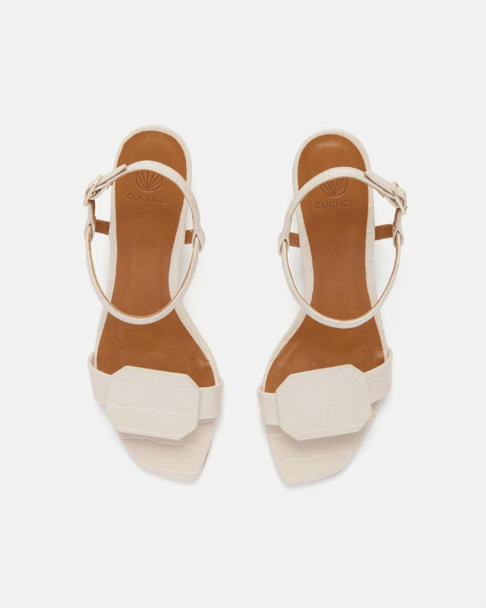 Front view of a bone white croc-embossed leather sandal with a slim ankle strap, octagonal toe detail, and a low wrapped demi heel on a white background.