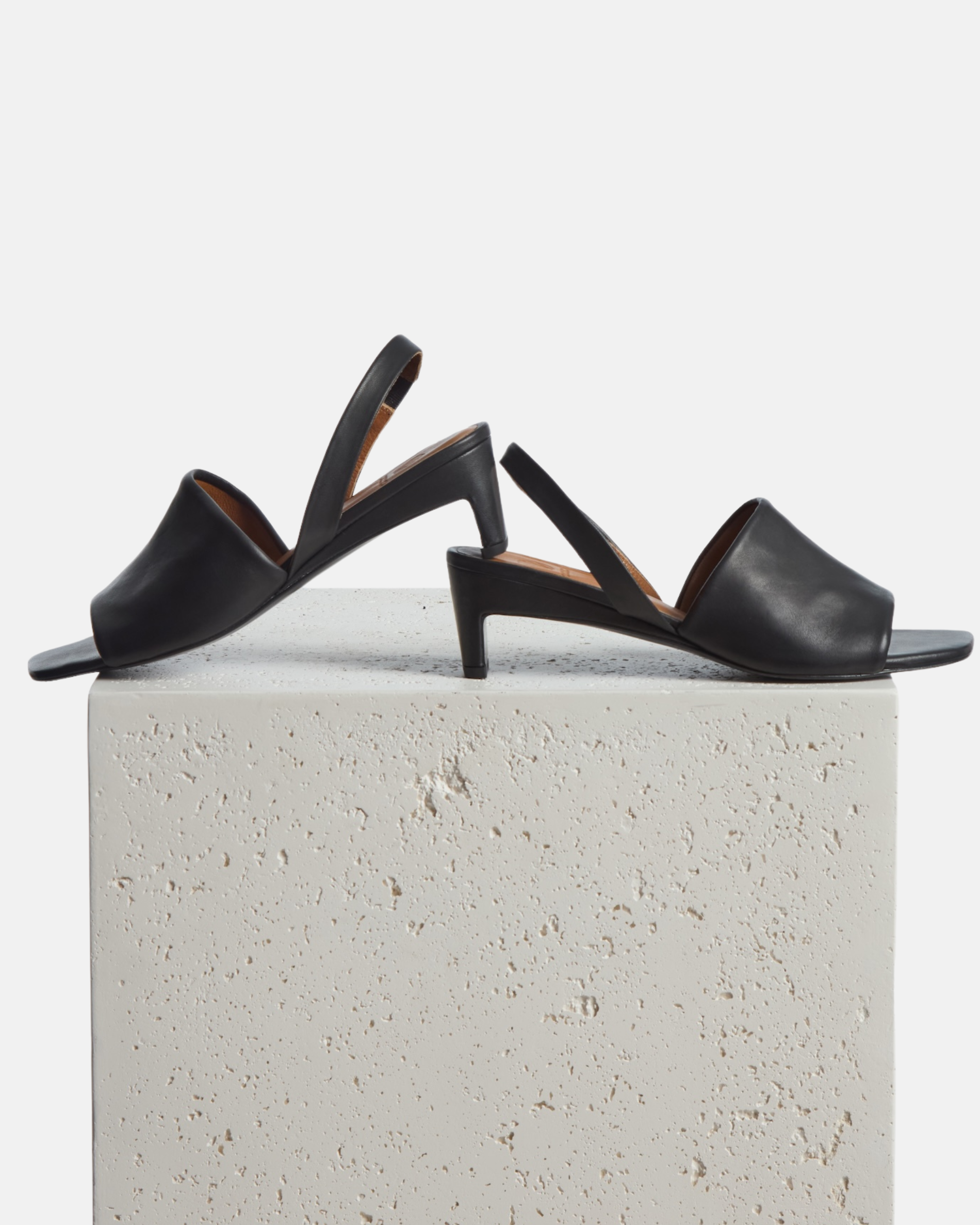 Shoes on a plinth: black leather mid-heel sandal with a wide band, delicate slingback, squared toe, and sculptural block heel