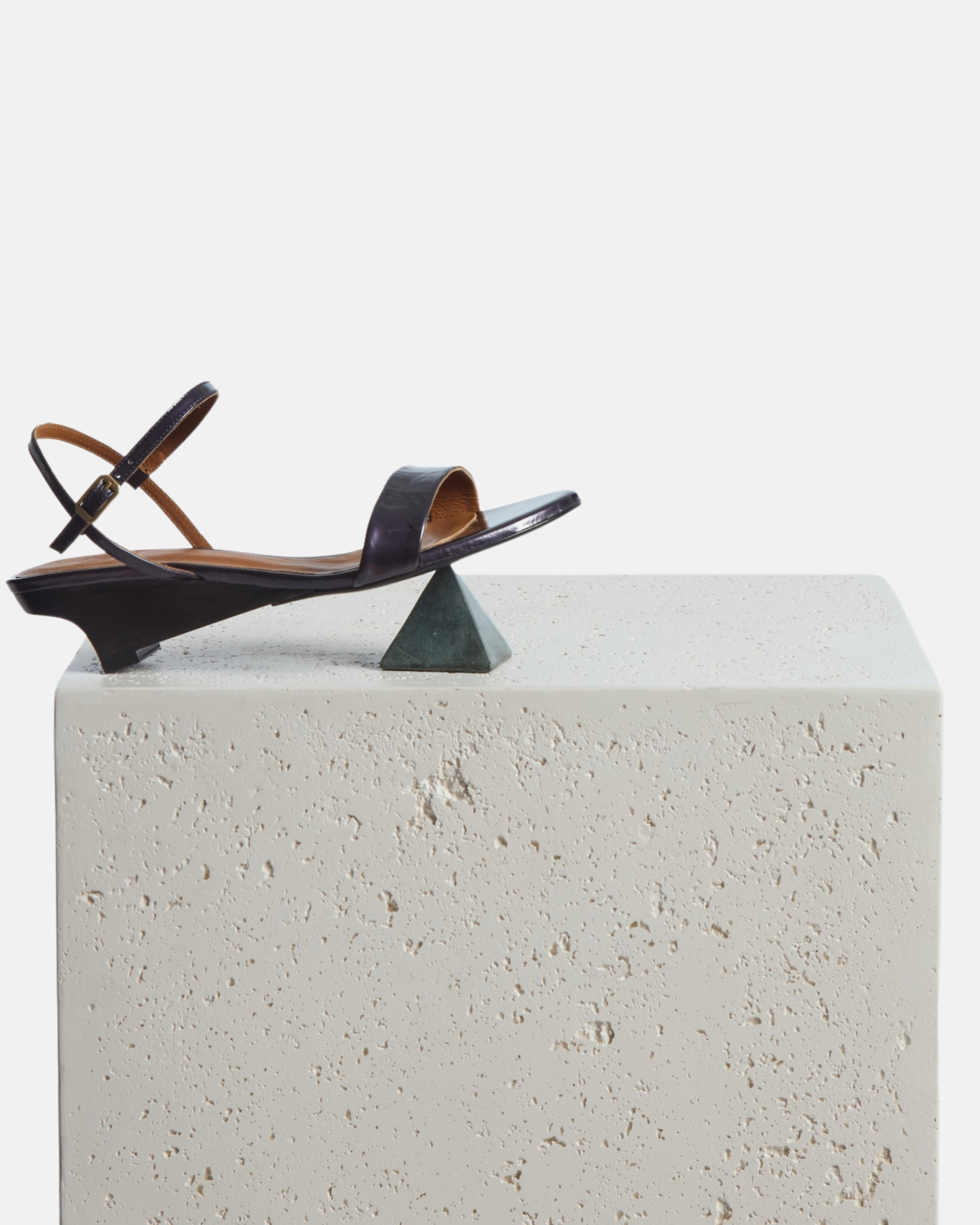 A shoe is perched on a plinth and a pyramid prop: black pearl patent leather sandal with a slim strap, ankle buckle, and sculptural wooden demi-wedge