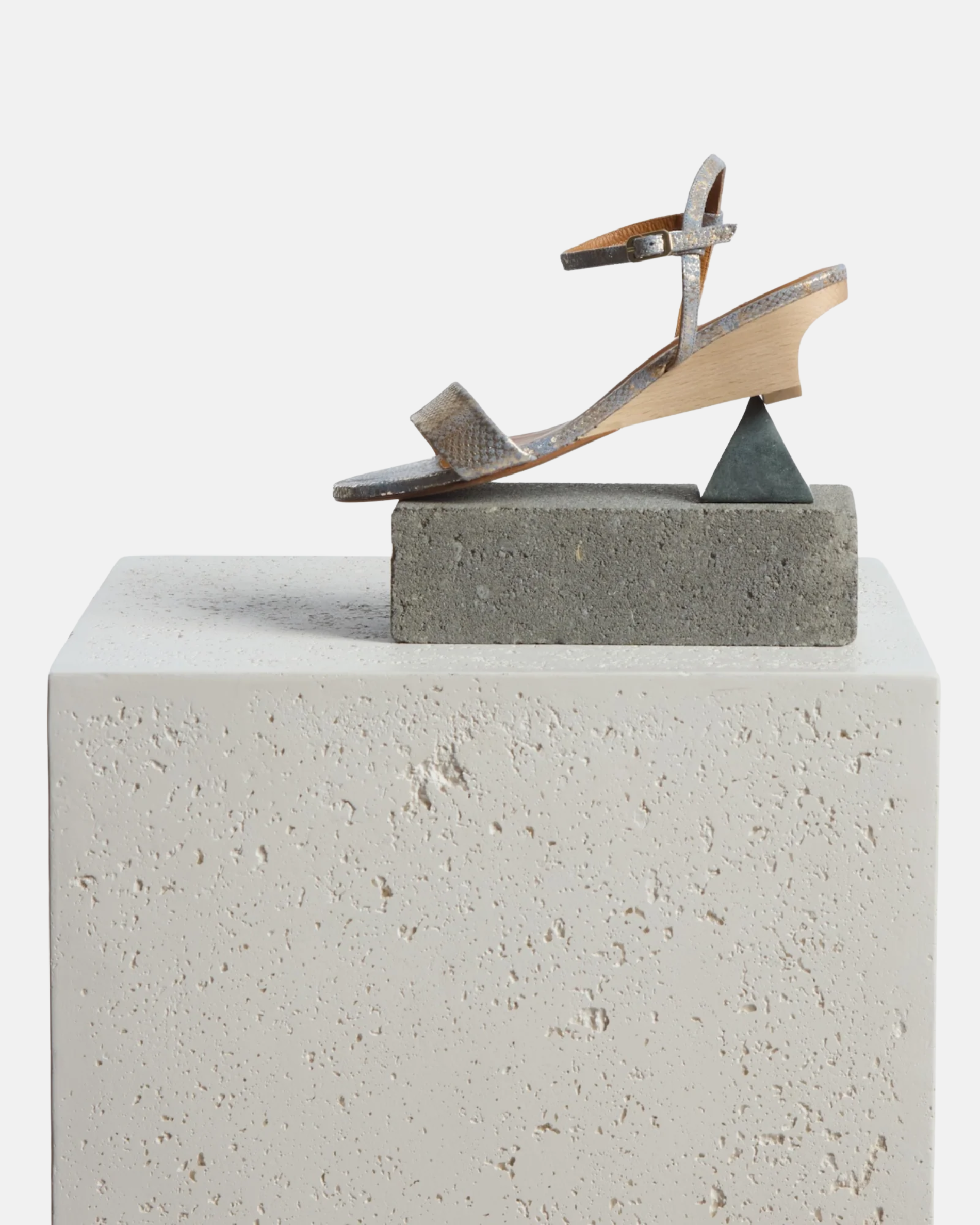 Shoe and objects placed on a plinth: gold and blue snake print leather demi-wedge sandal with a slim strap, ankle buckle, and sculptural wooden wedge.