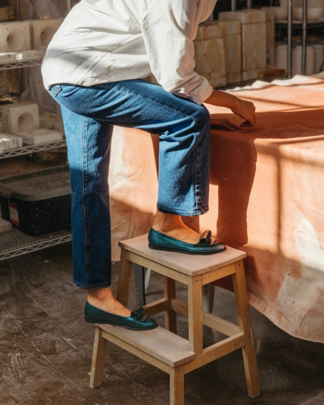 A person wearing blue jeans, a white shirt, and teal loafers is standing on a small wooden step stool next to a table covered with a peach-colored cloth.