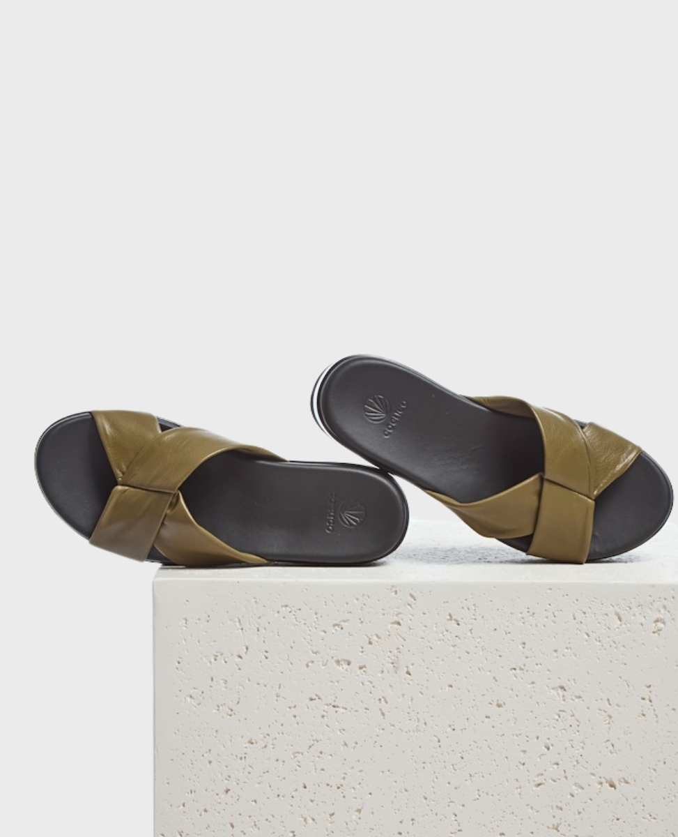 A pair of olive green leather sandals with crisscross straps, a smooth black footbed, and a minimalist design, resting on a textured white surface.