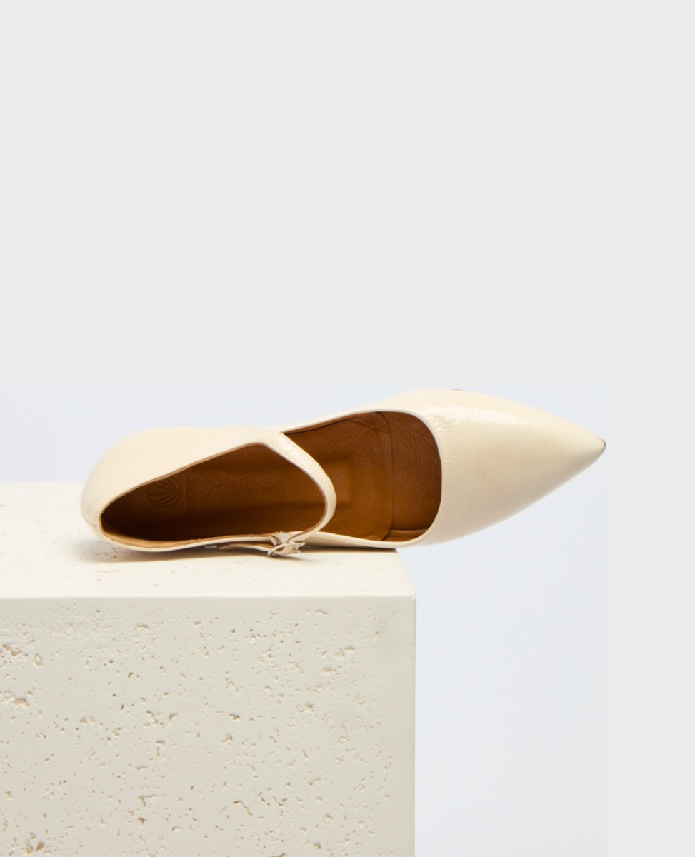 A sleek, pointed-toe, cream-colored flat shoe with a smooth, glossy finish and a thin ankle strap, resting on a textured, light-colored block.