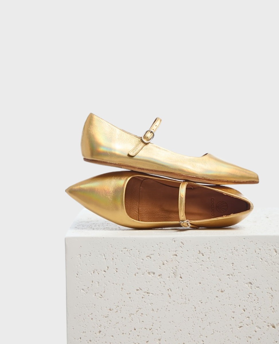 A pair of sleek, pointed-toe flats in a shimmering gold color with a smooth, metallic finish, featuring a thin strap with a small buckle across the top, resting on a textured white surface.